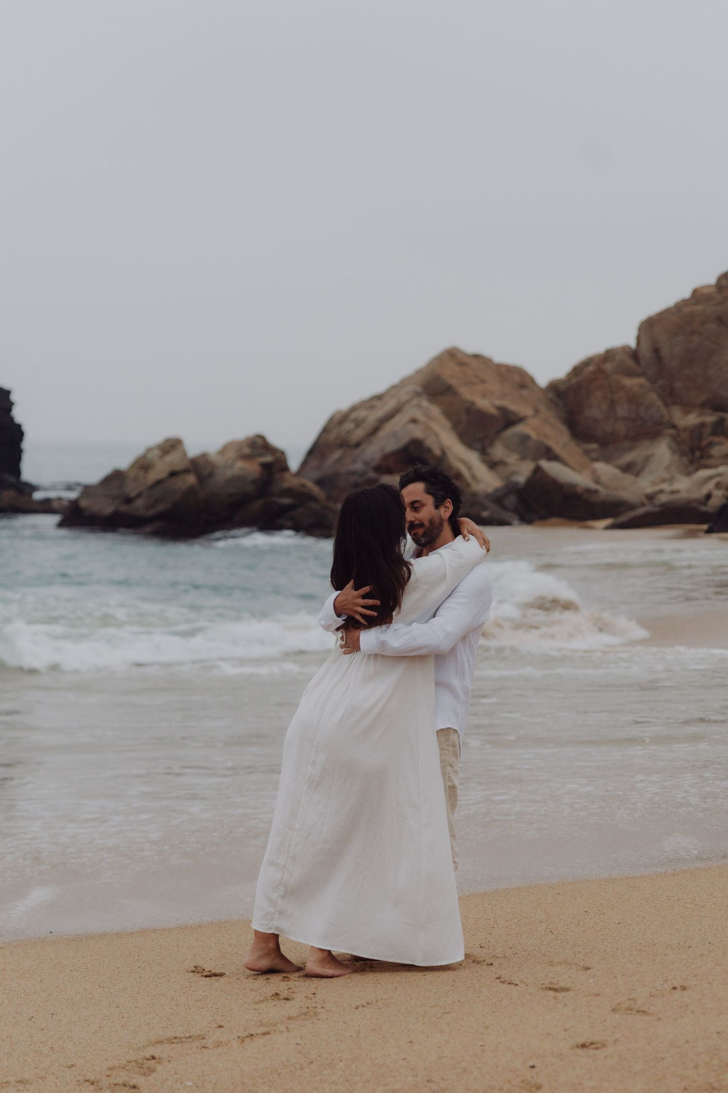 Romantic couple embracing on sandy beach in white outfits with rocky coastline and ocean waves behind them.