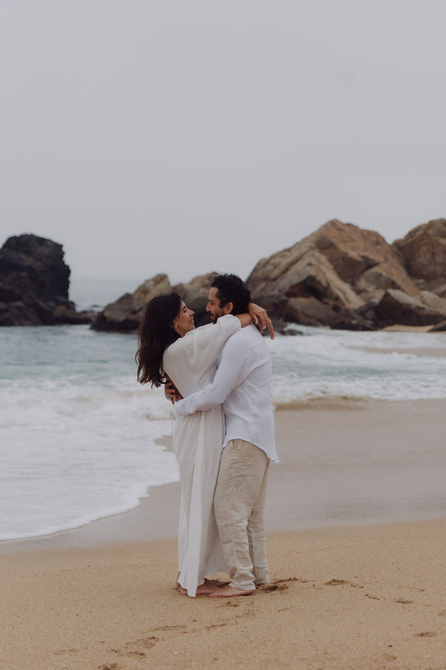 Romantic couple in white outfits embracing on sandy beach with rocky coastline and ocean waves behind them.