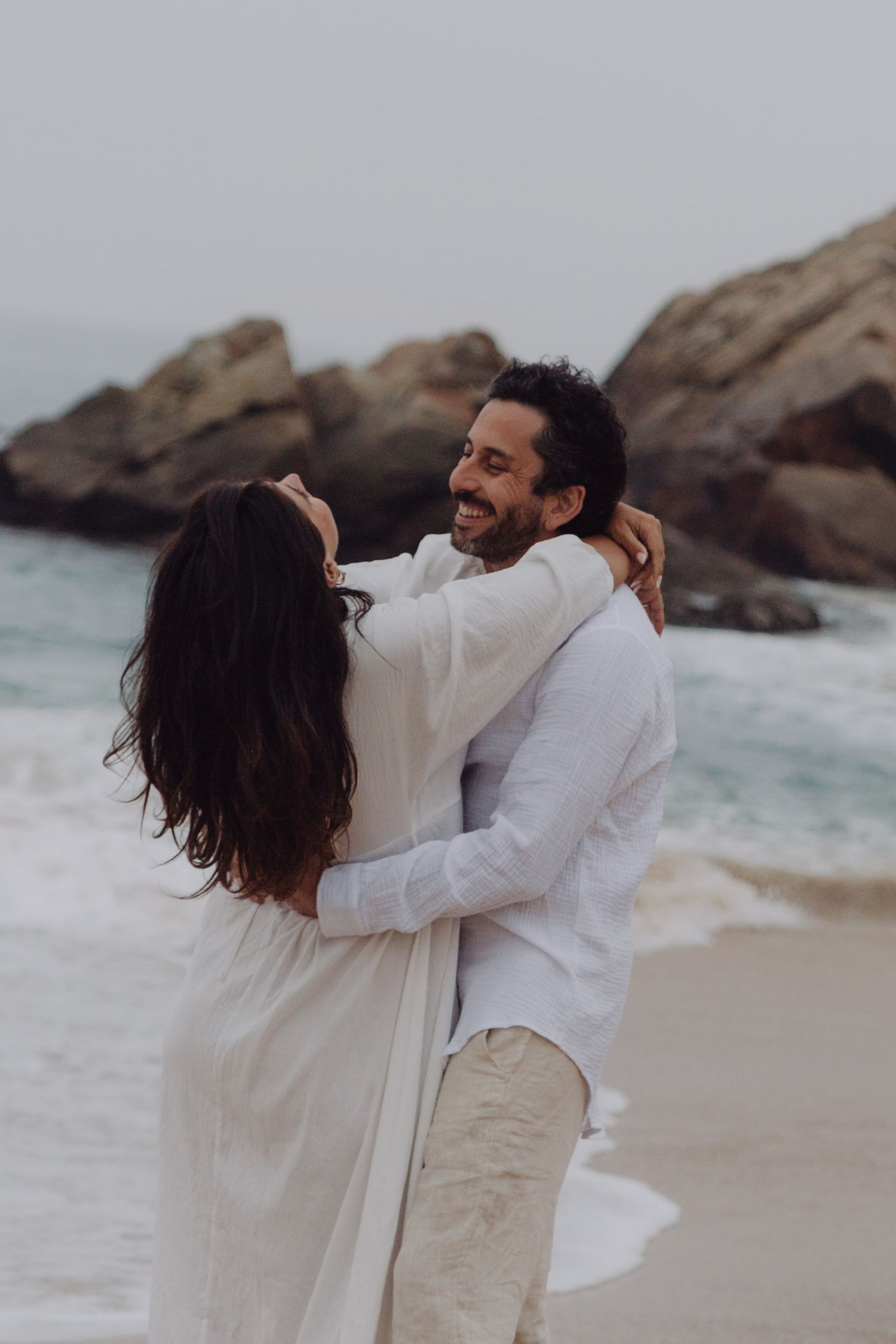 Smiling man in white shirt embracing woman in white dress on sandy beach with rocky ocean backdrop.