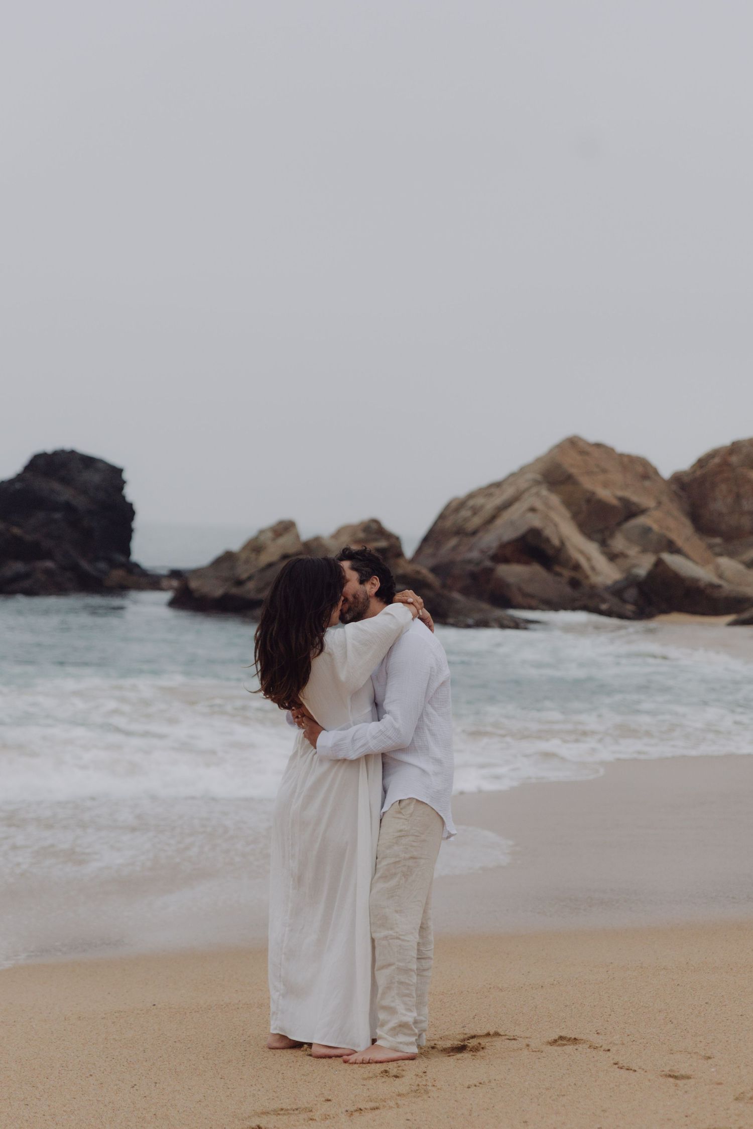 Couple sharing a kiss on sandy beach in white outfits with rocky coastline and ocean waves behind them.