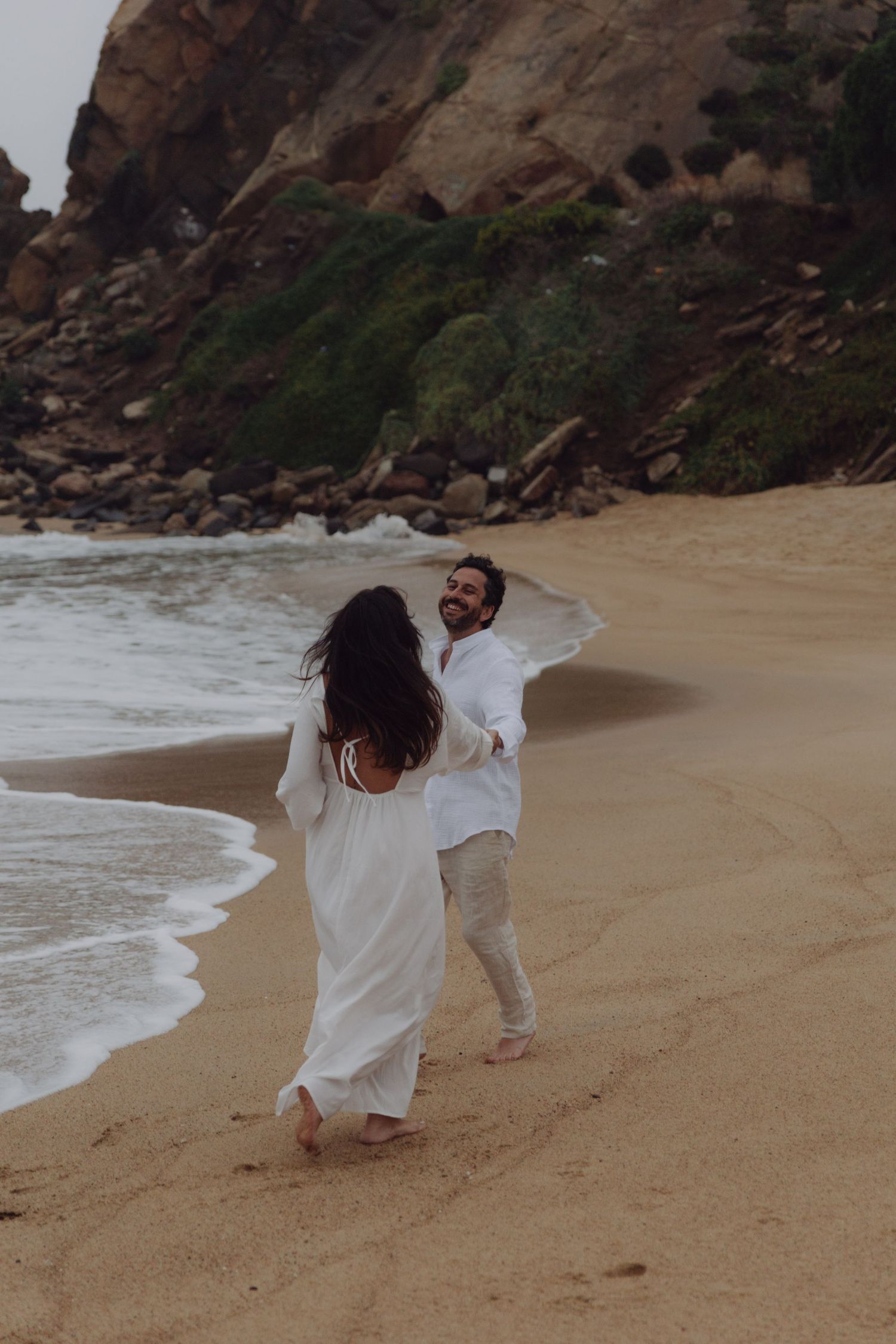 Joyful couple in white outfits running and playing on sandy beach with rocky cliffs and ocean waves.