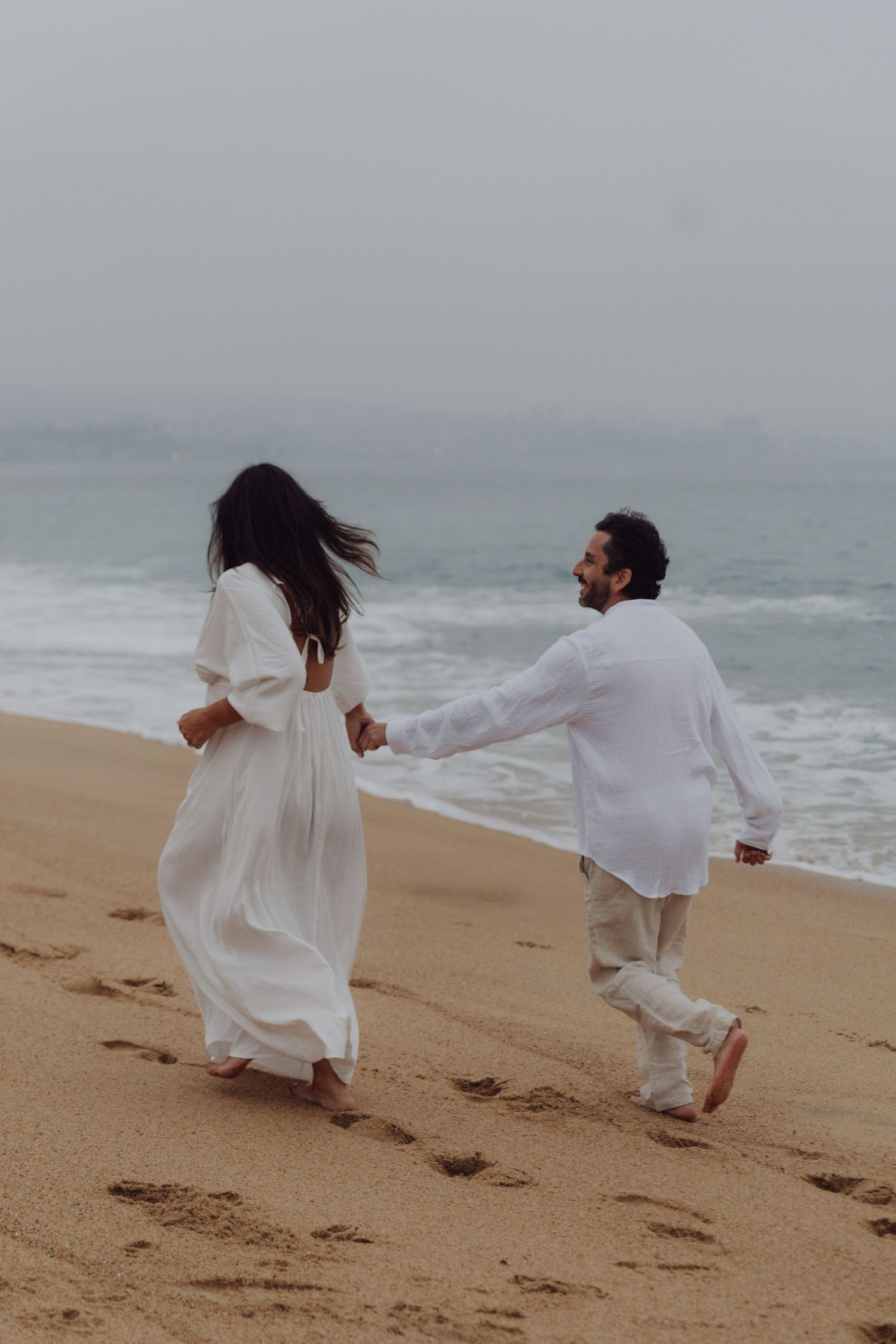 Couple in white outfits holding hands and walking barefoot on a sandy beach with ocean waves.