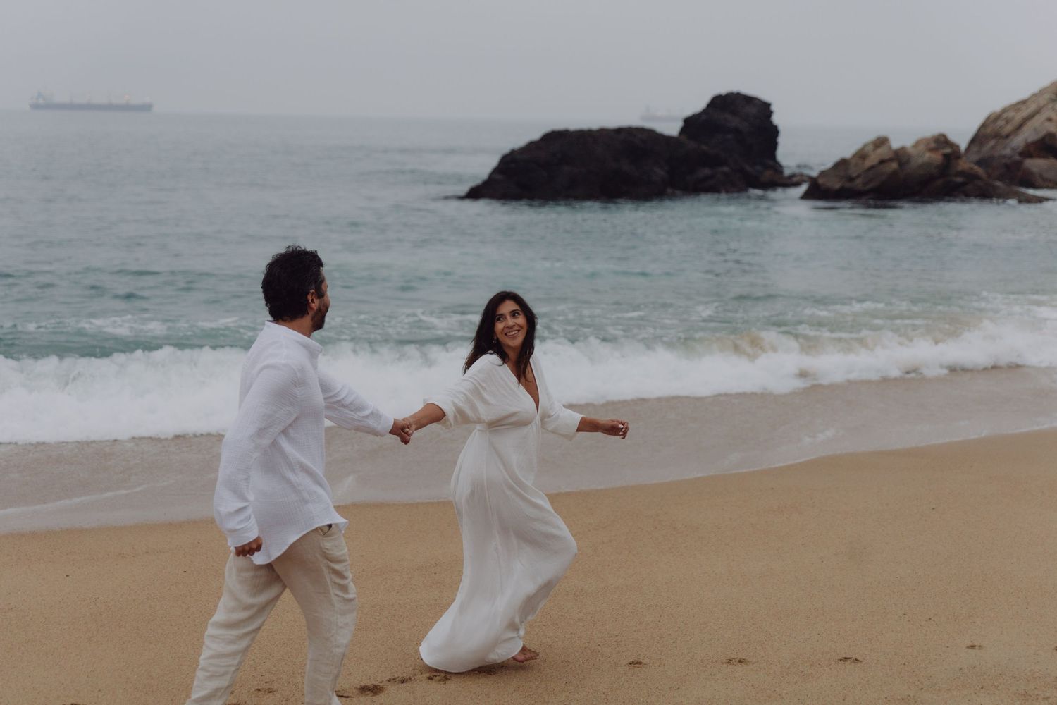 Romantic couple dressed in white holding hands on a rocky beach with ocean backdrop.