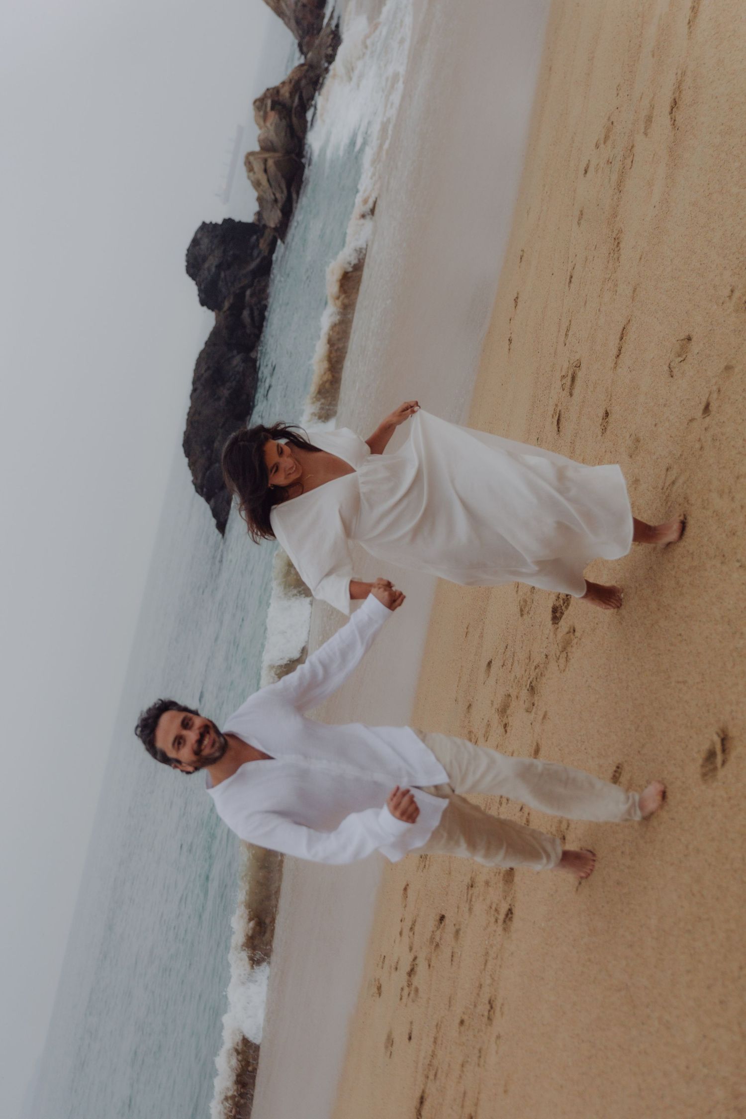 Joyful couple in matching white outfits running together on a sandy beach near rocks.