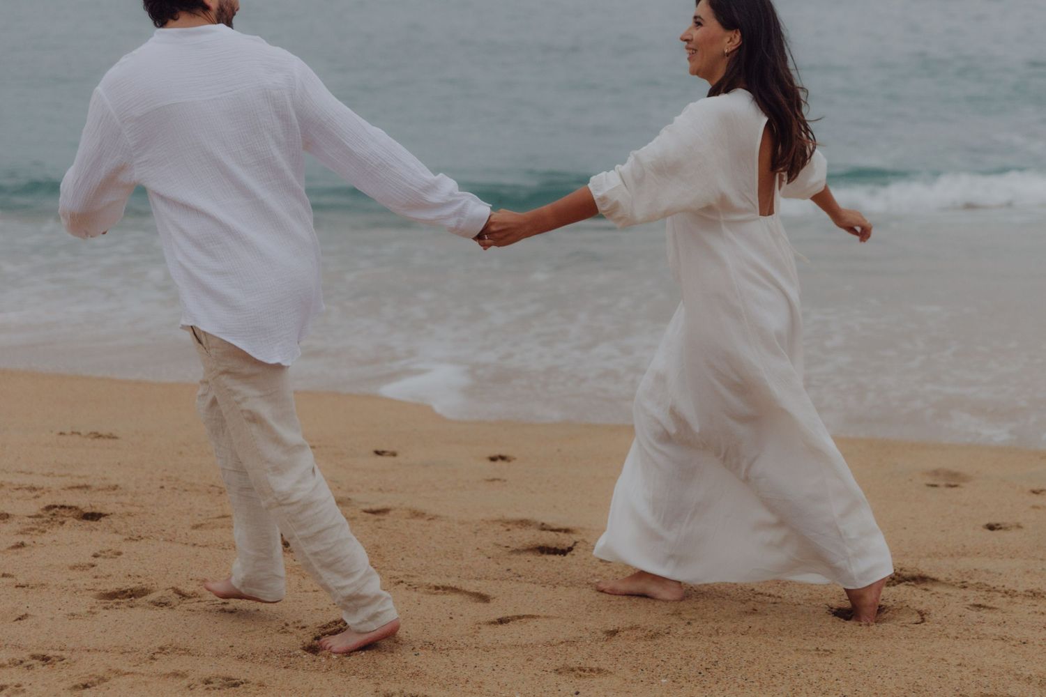 Couple in white clothing holding hands and walking along a sandy beach by the ocean.
