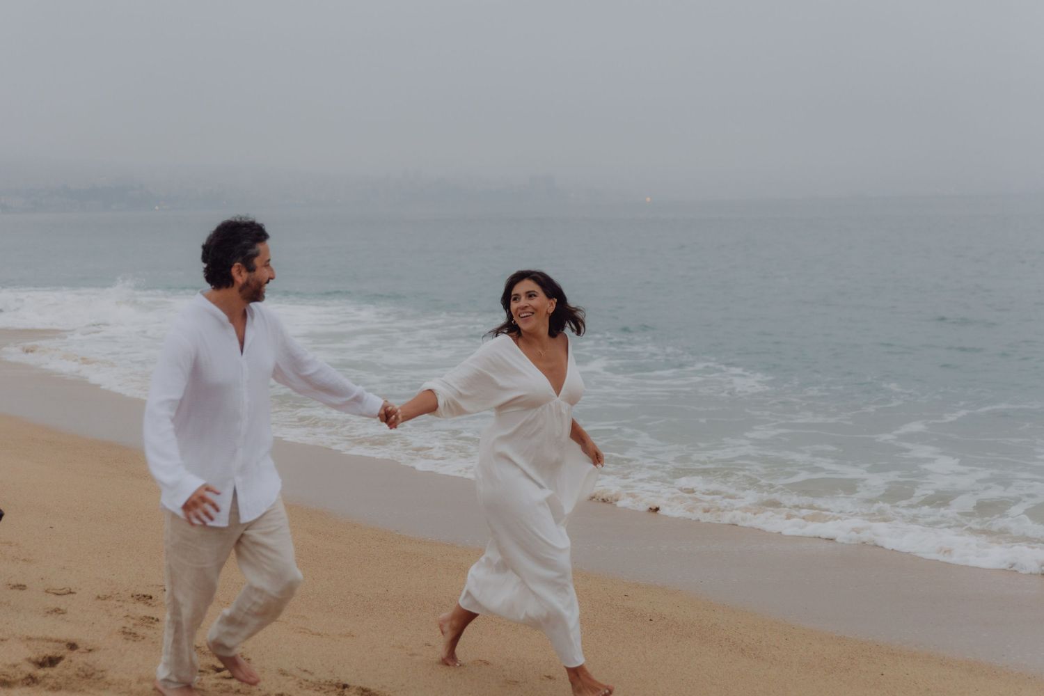 Happy couple in white outfits laughing and running together on a misty beach shoreline.