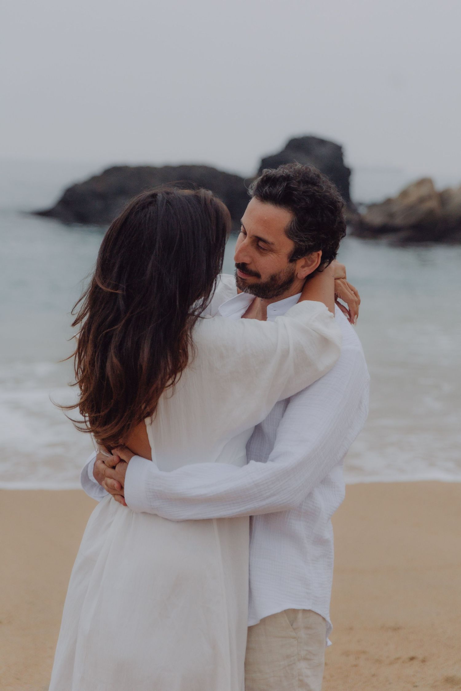 Romantic couple in white embracing on a sandy beach with rocky ocean backdrop.