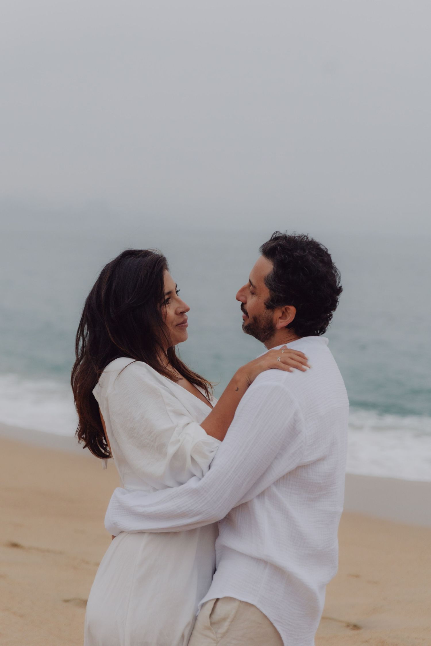 Smiling couple in white outfits embracing and gazing at each other on a beach.