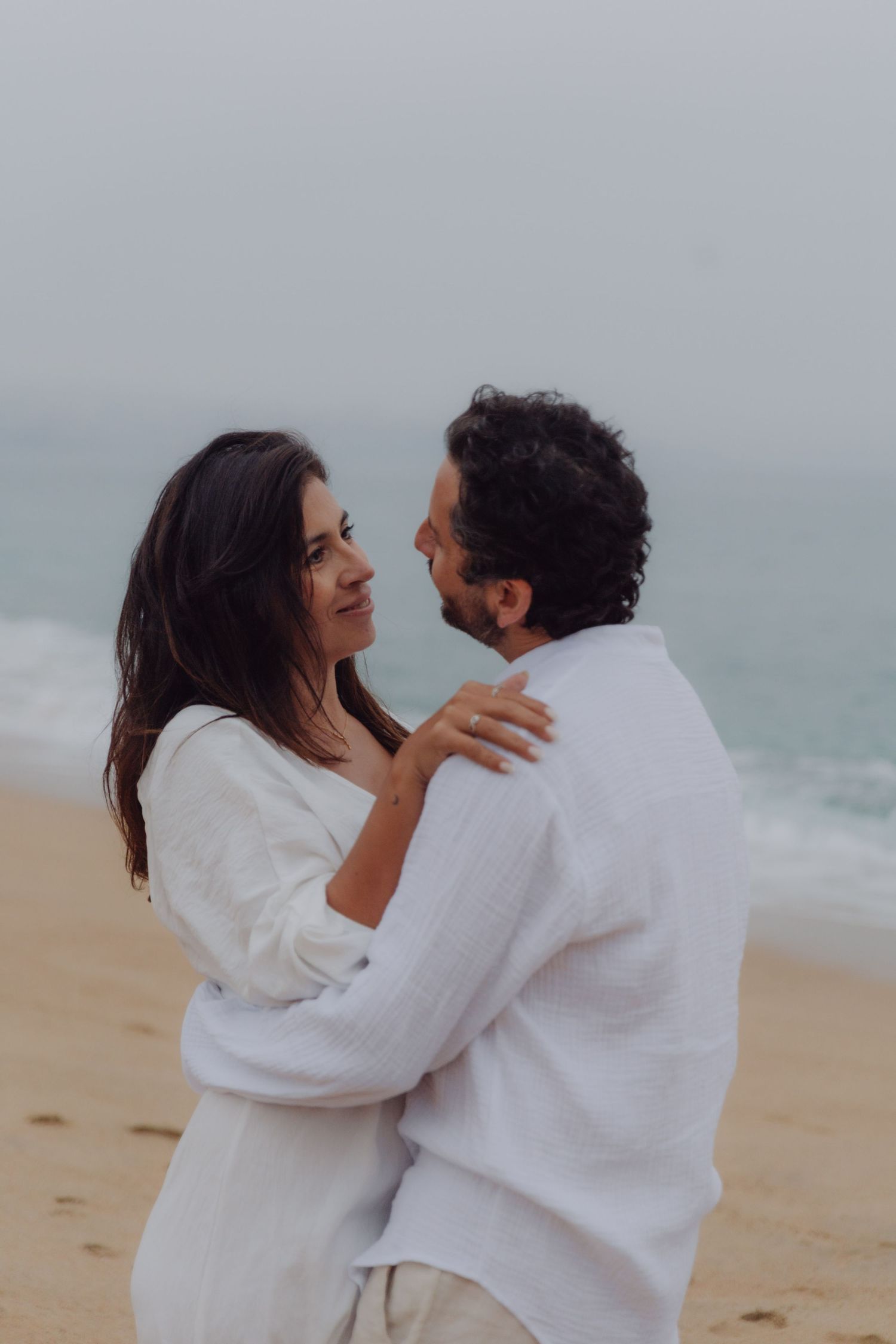 Loving couple in white holding each other close on a sandy beach with ocean waves.