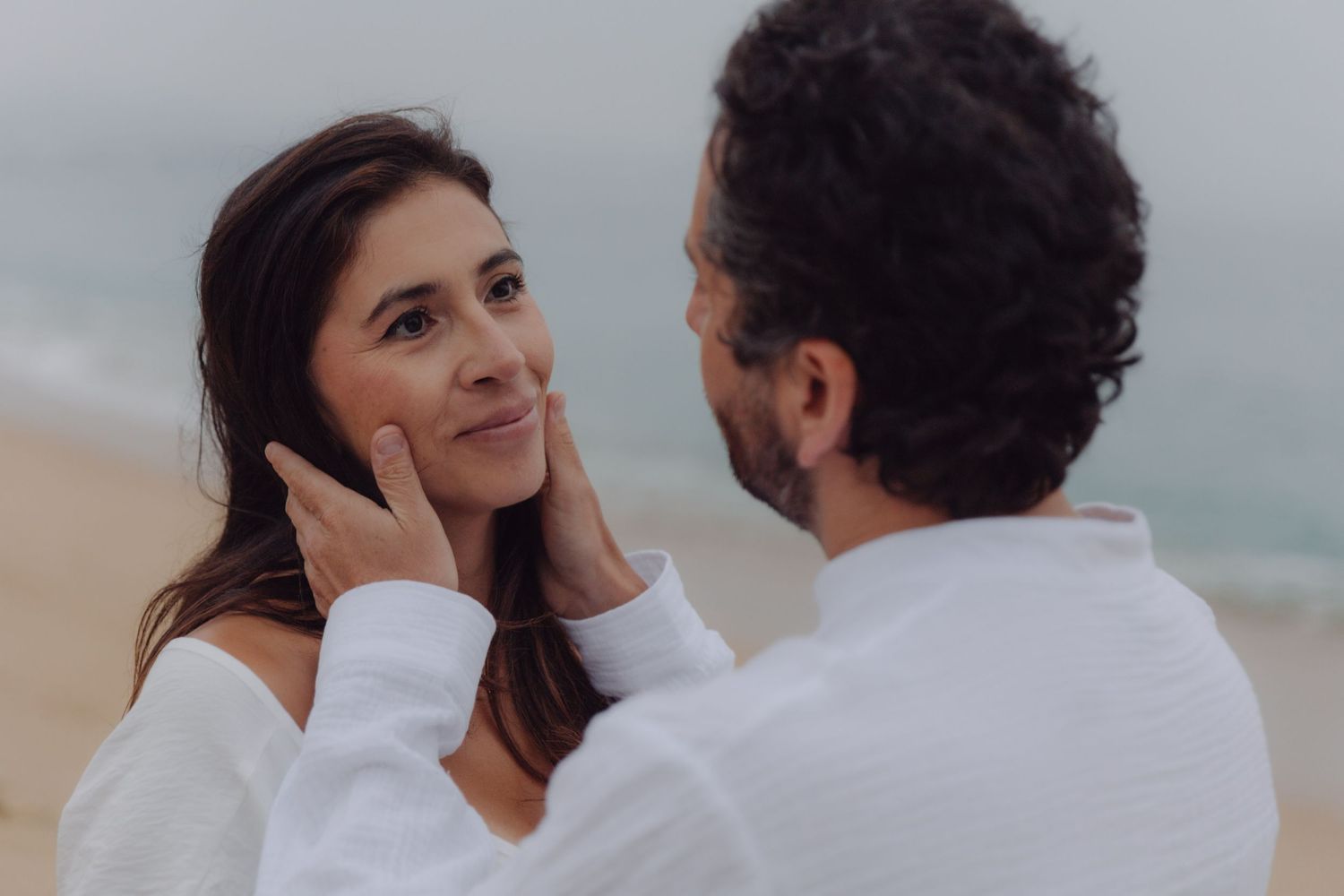 Man tenderly touching womans face as she smiles at him on a misty beach.
