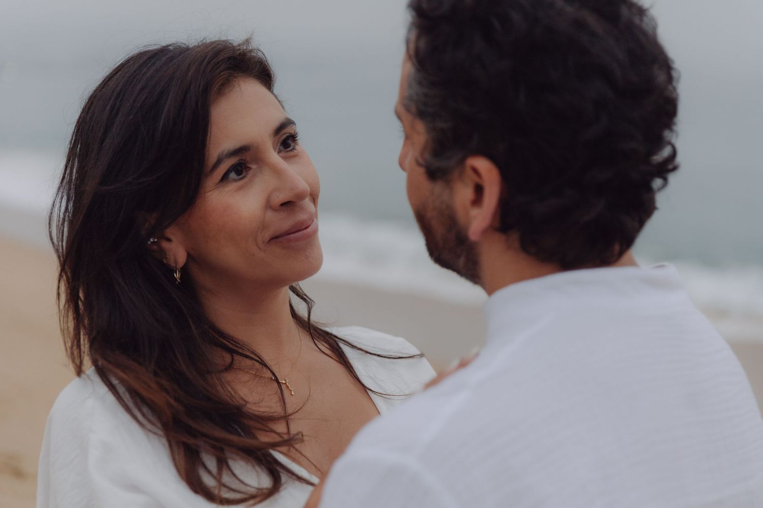 Close-up of a woman smiling at her partner on a breezy beach with ocean behind.