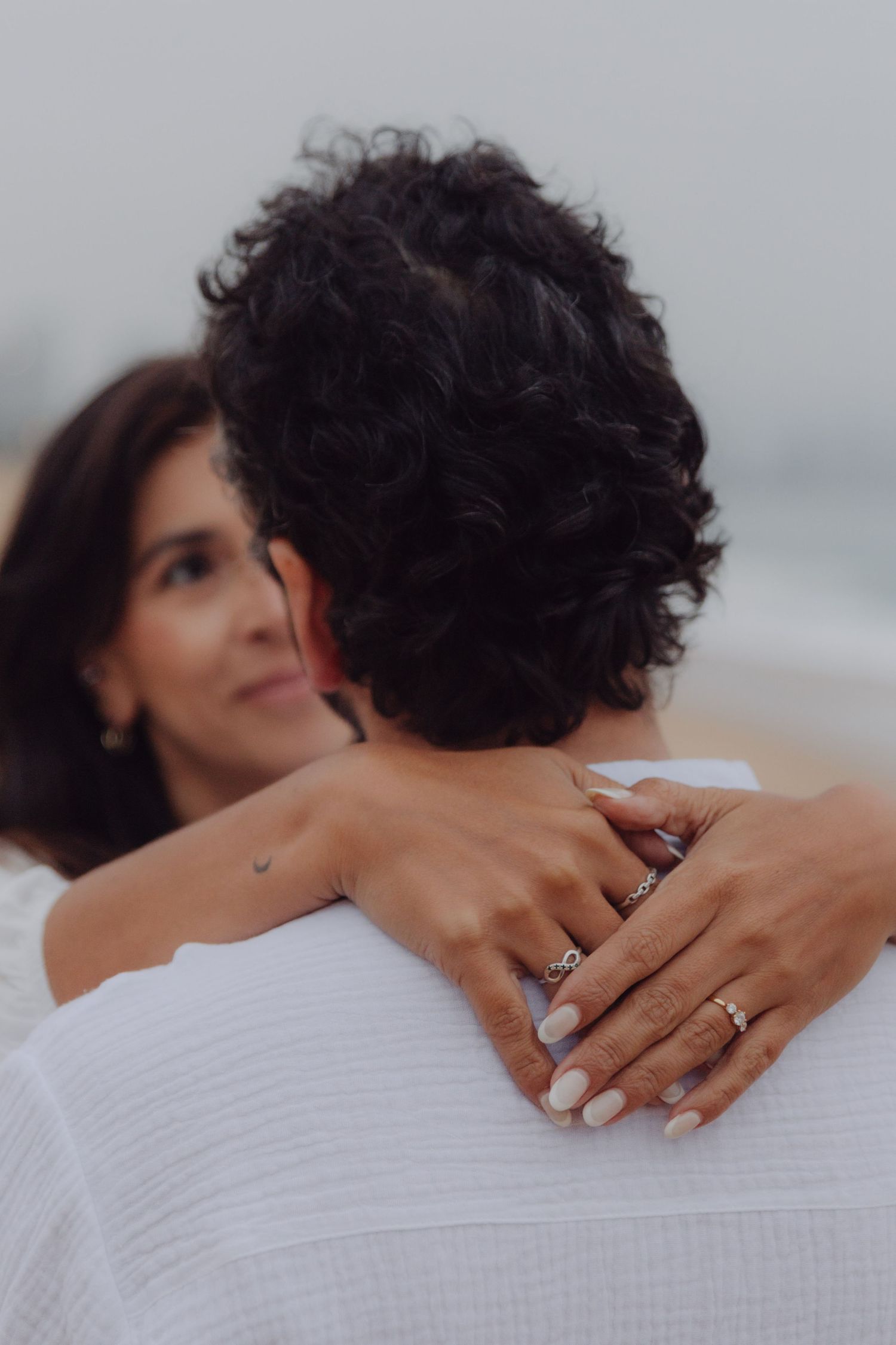 Couple embracing on beach, woman's hands with rings wrapped around man's shoulders in romantic moment.