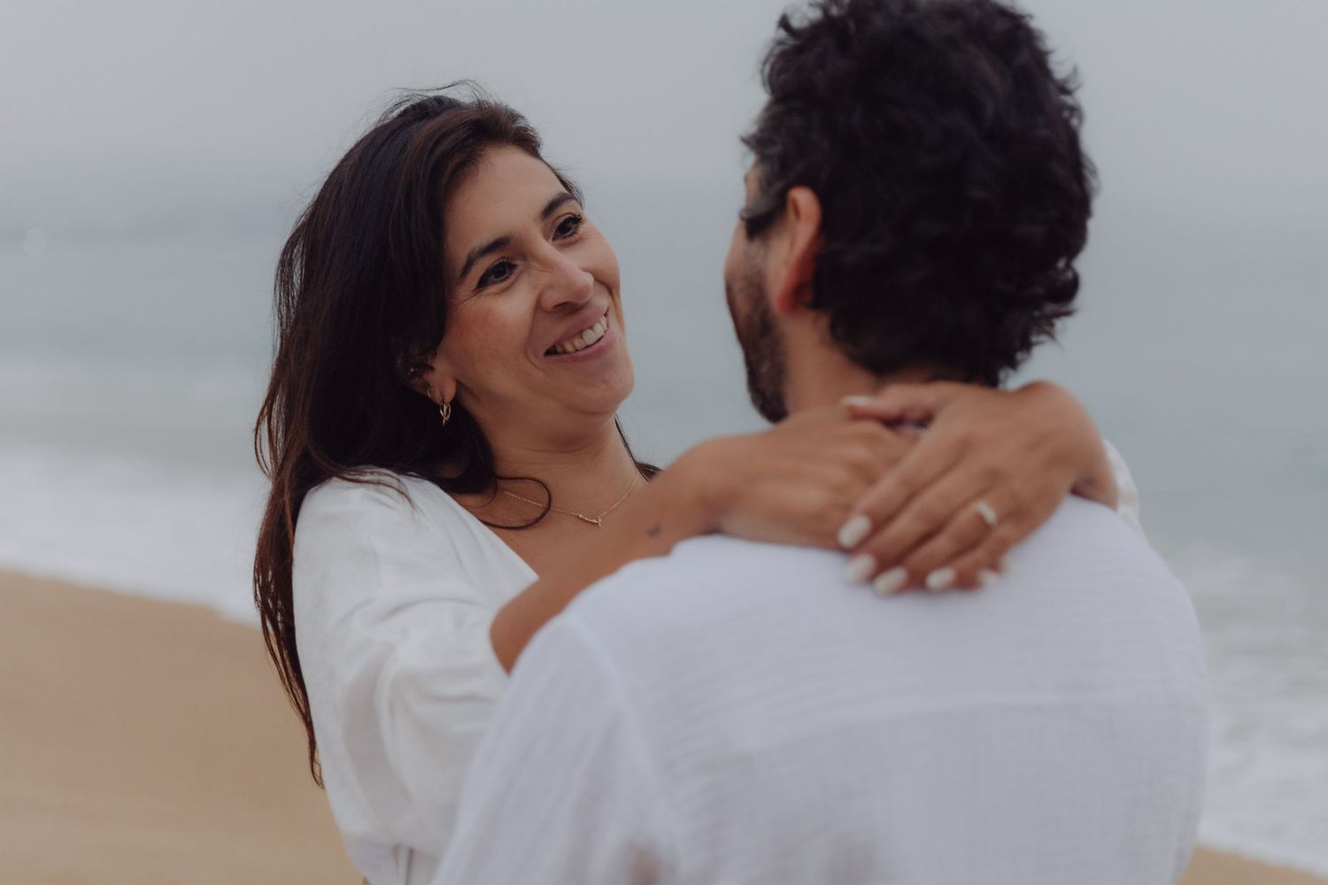 Smiling woman in white outfit embraces man on sandy beach with ocean waves in background.
