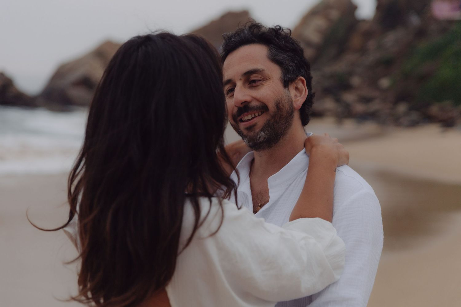 Bearded man in white shirt smiles warmly at woman during romantic beach photo session.