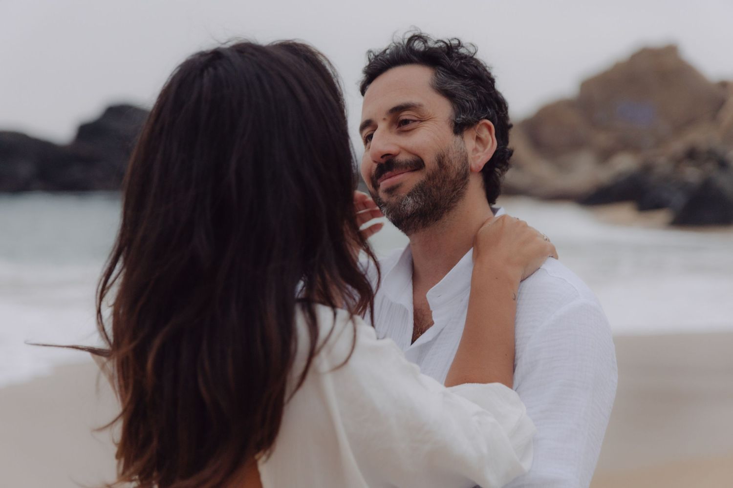 Happy bearded man in white shirt shares tender moment with woman on scenic beach.