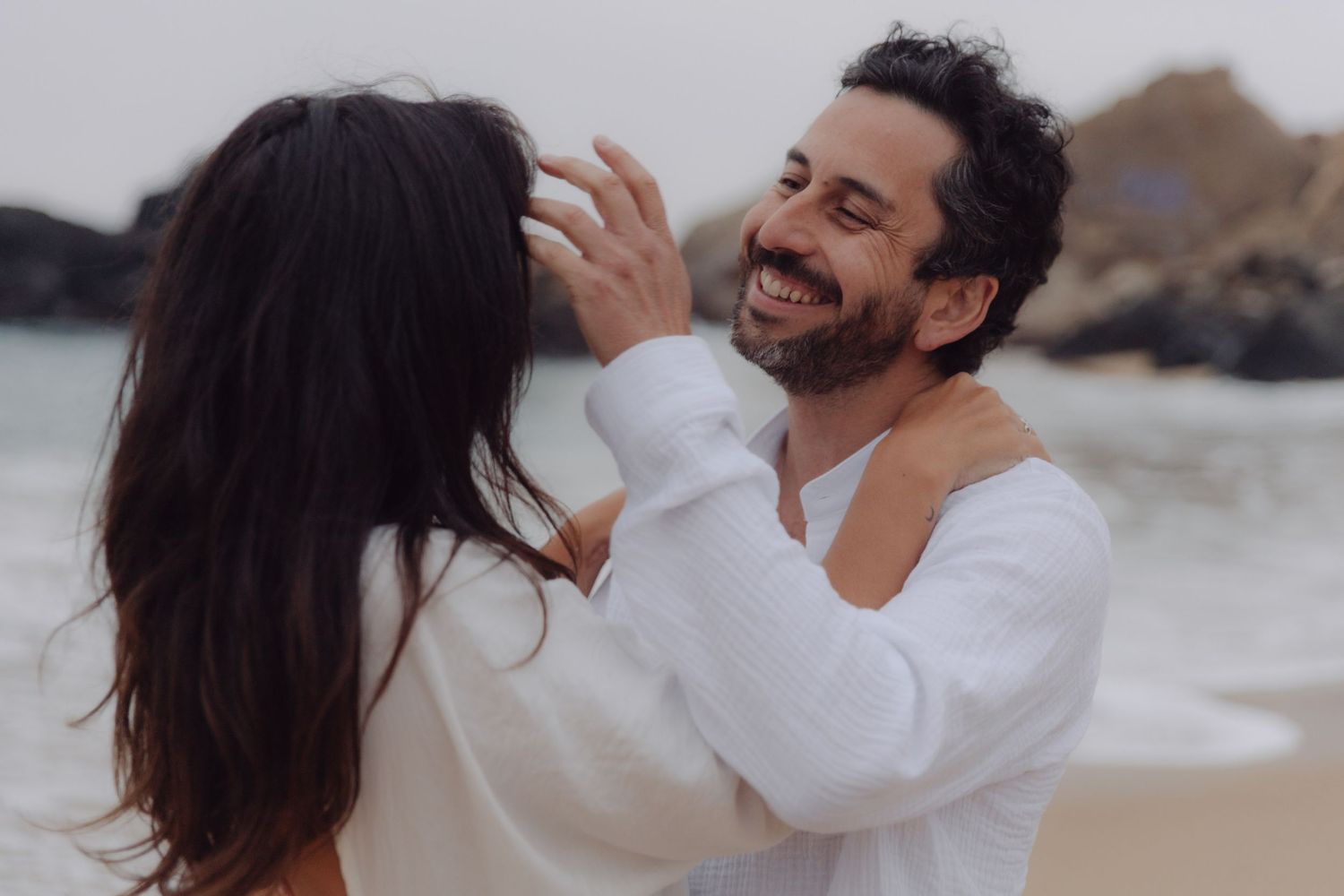 Smiling bearded man gently touches woman's hair during romantic couples photoshoot on beach.