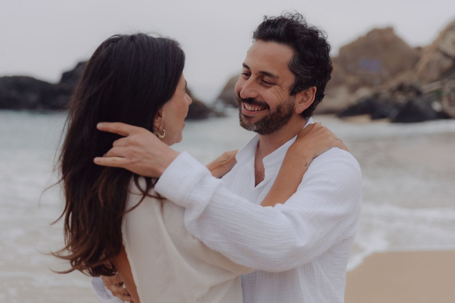 Joyful couple in white outfits share a loving gaze on a rocky beach shoreline.
