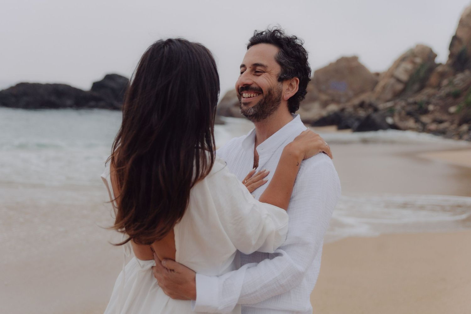 Laughing couple in white outfits embrace each other on a sandy beach with rocks.