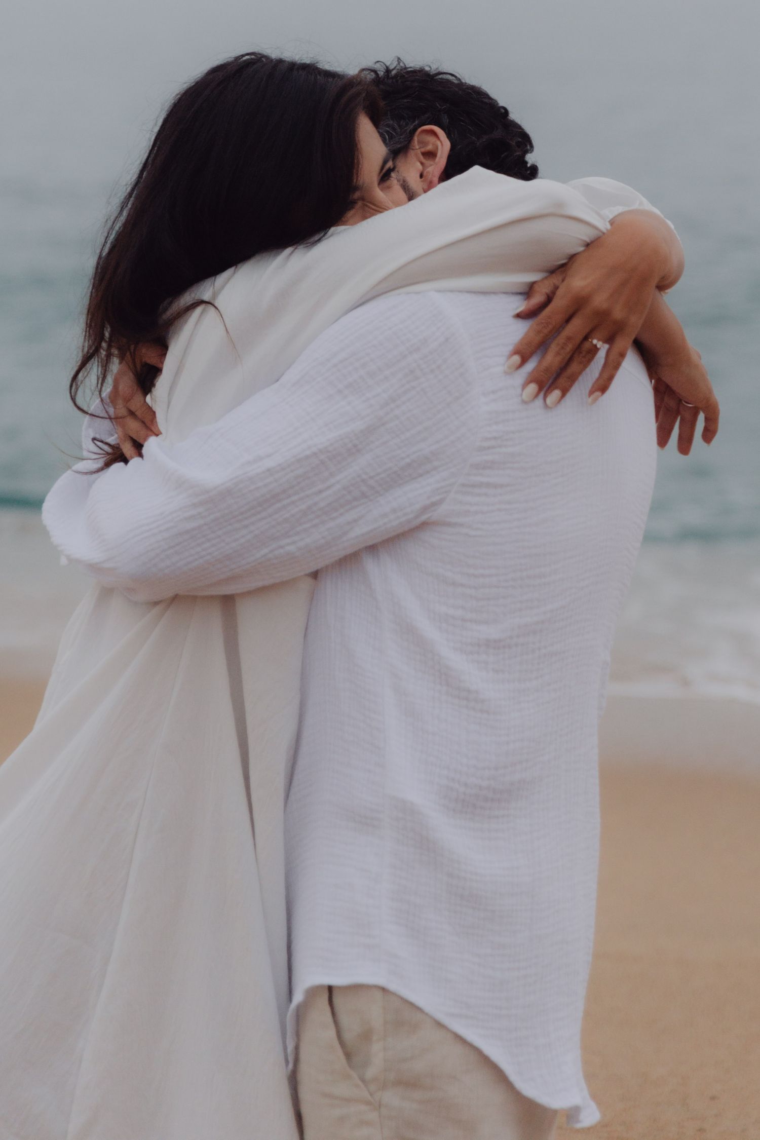 Couple in matching white outfits share a warm embrace on a sandy ocean beach.