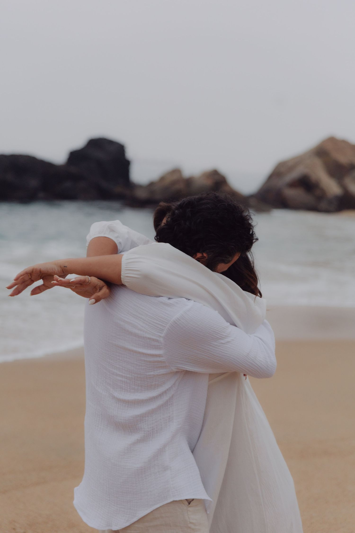 Couple in white clothing share a tender embrace on a sandy beach with rock formations.