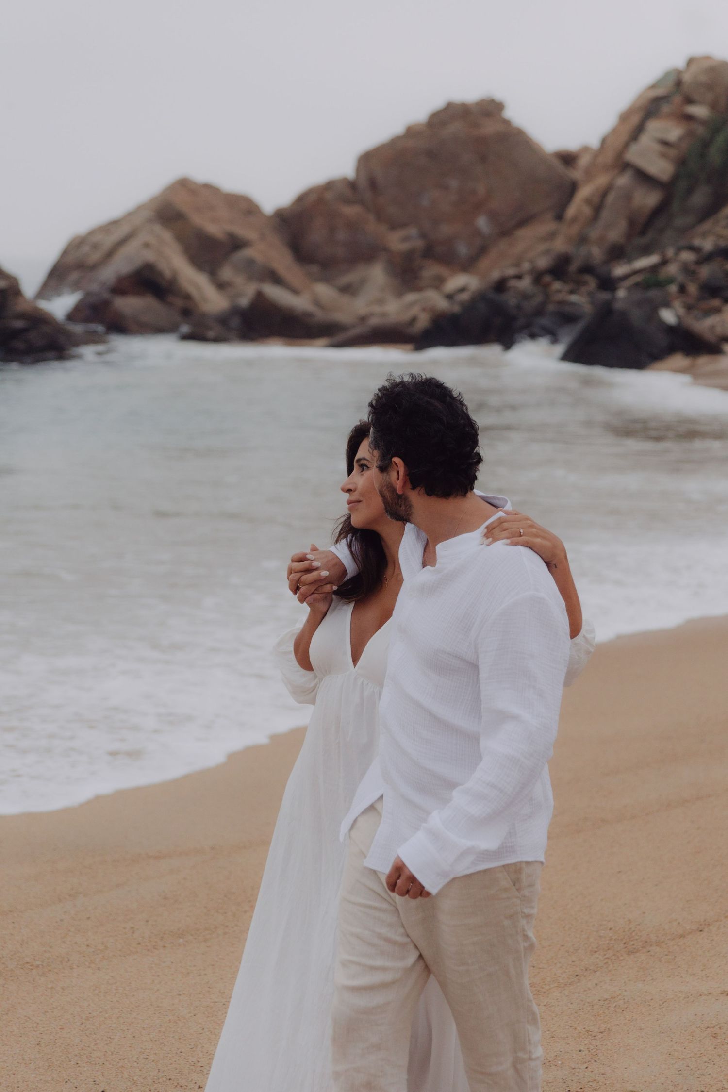 Romantic couple in white outfits stand together on sandy beach with dramatic rock backdrop.