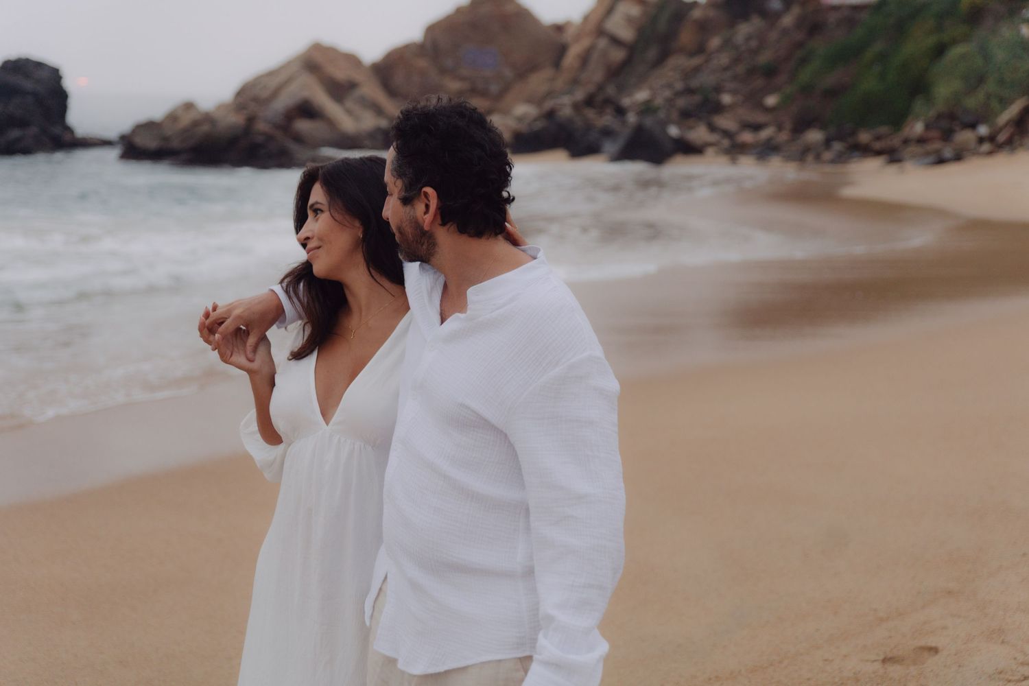 Couple in white outfits walking on a sandy beach with rocky cliffs in the background.