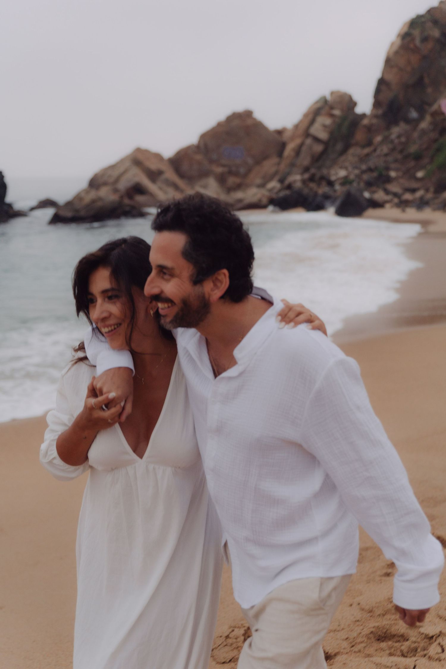 Smiling couple in white linen embracing on a beach with ocean waves and rocks behind them.