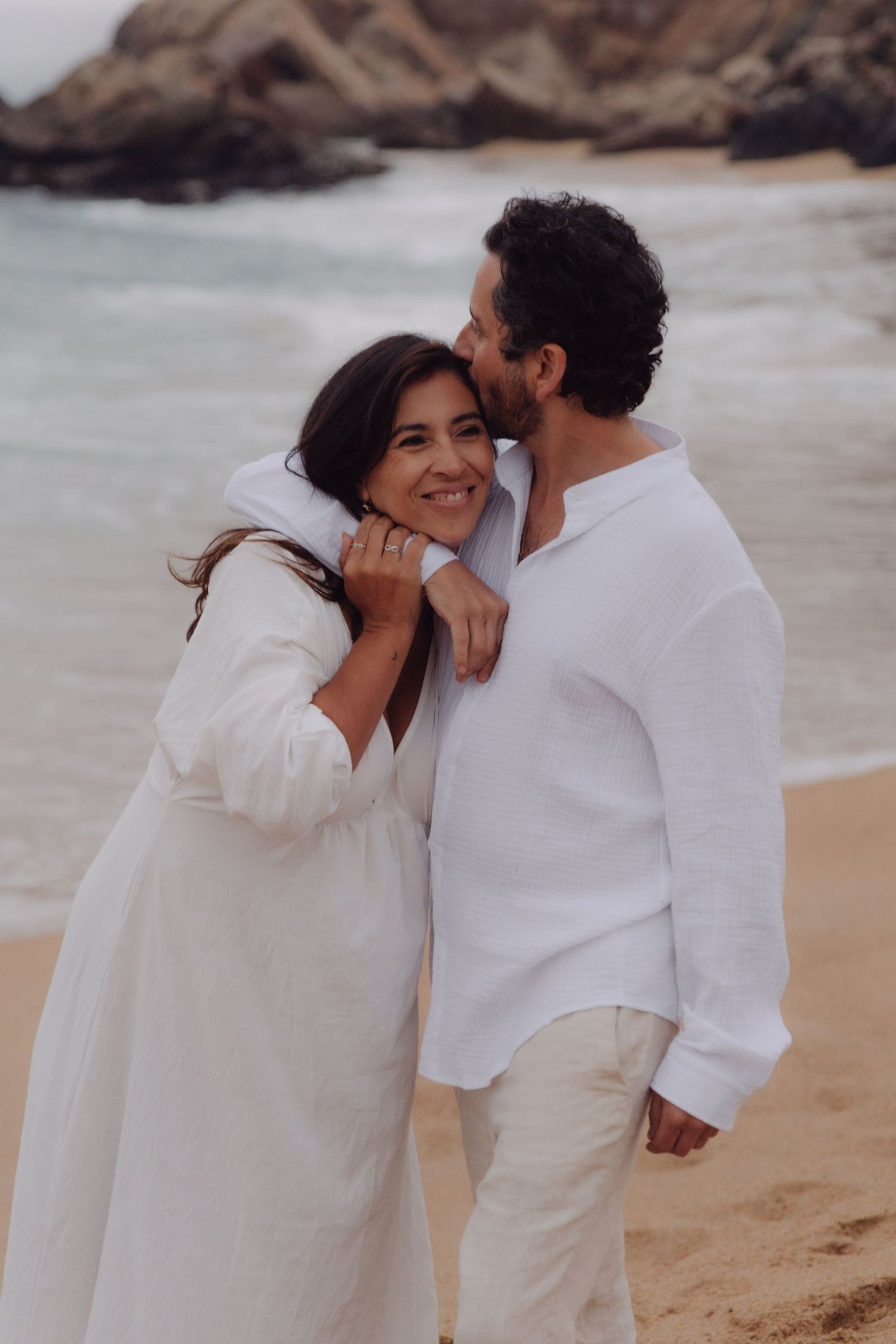 Man kissing woman on cheek while embracing on beach, both wearing white, woman smiling with ring visible.