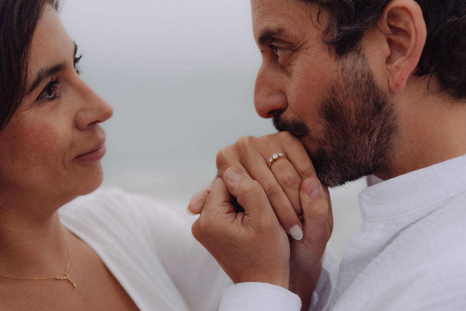 Close-up of man kissing woman's hand showing engagement ring on a misty beach backdrop.