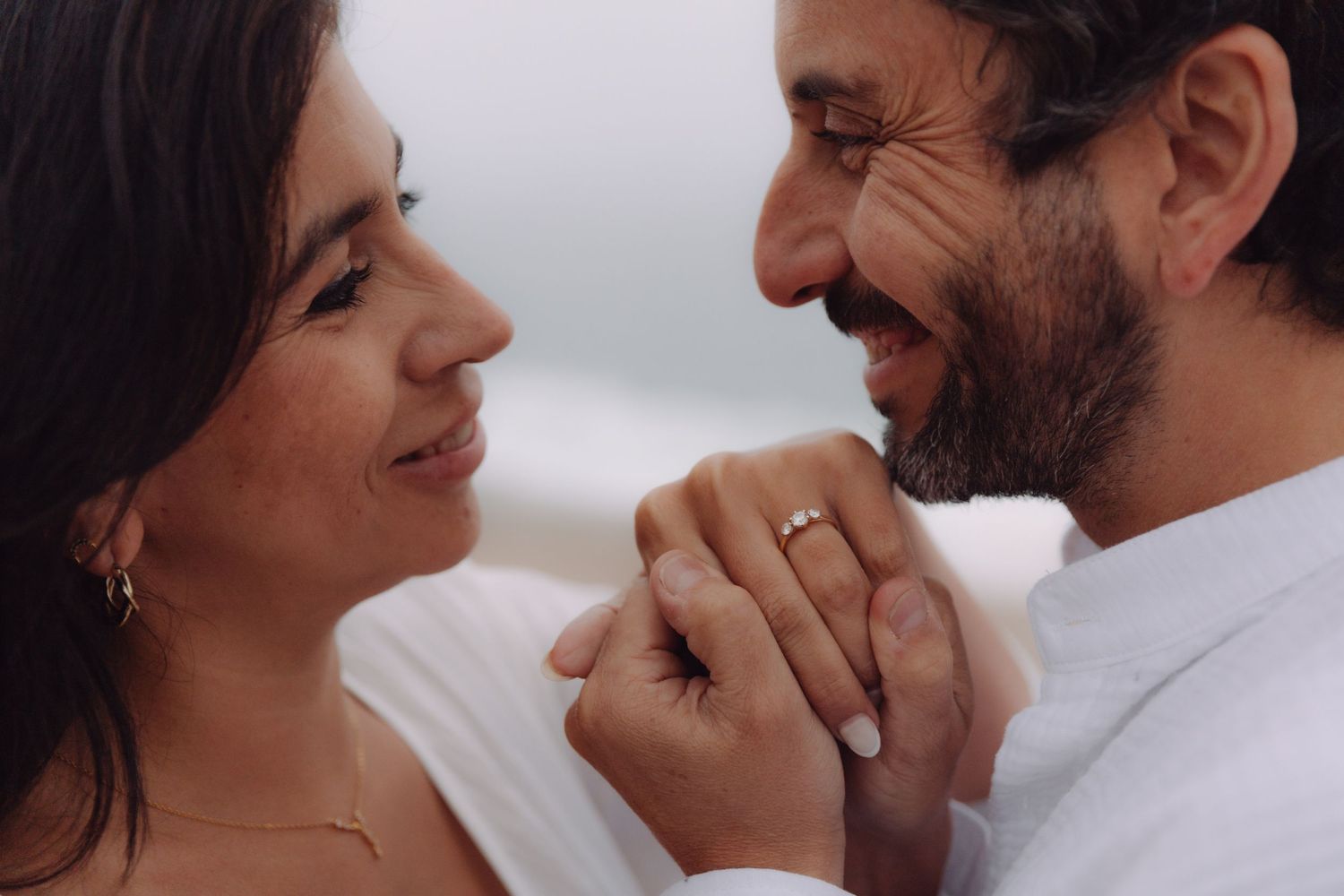 Couple smiling face-to-face holding hands with engagement ring visible against a soft background.