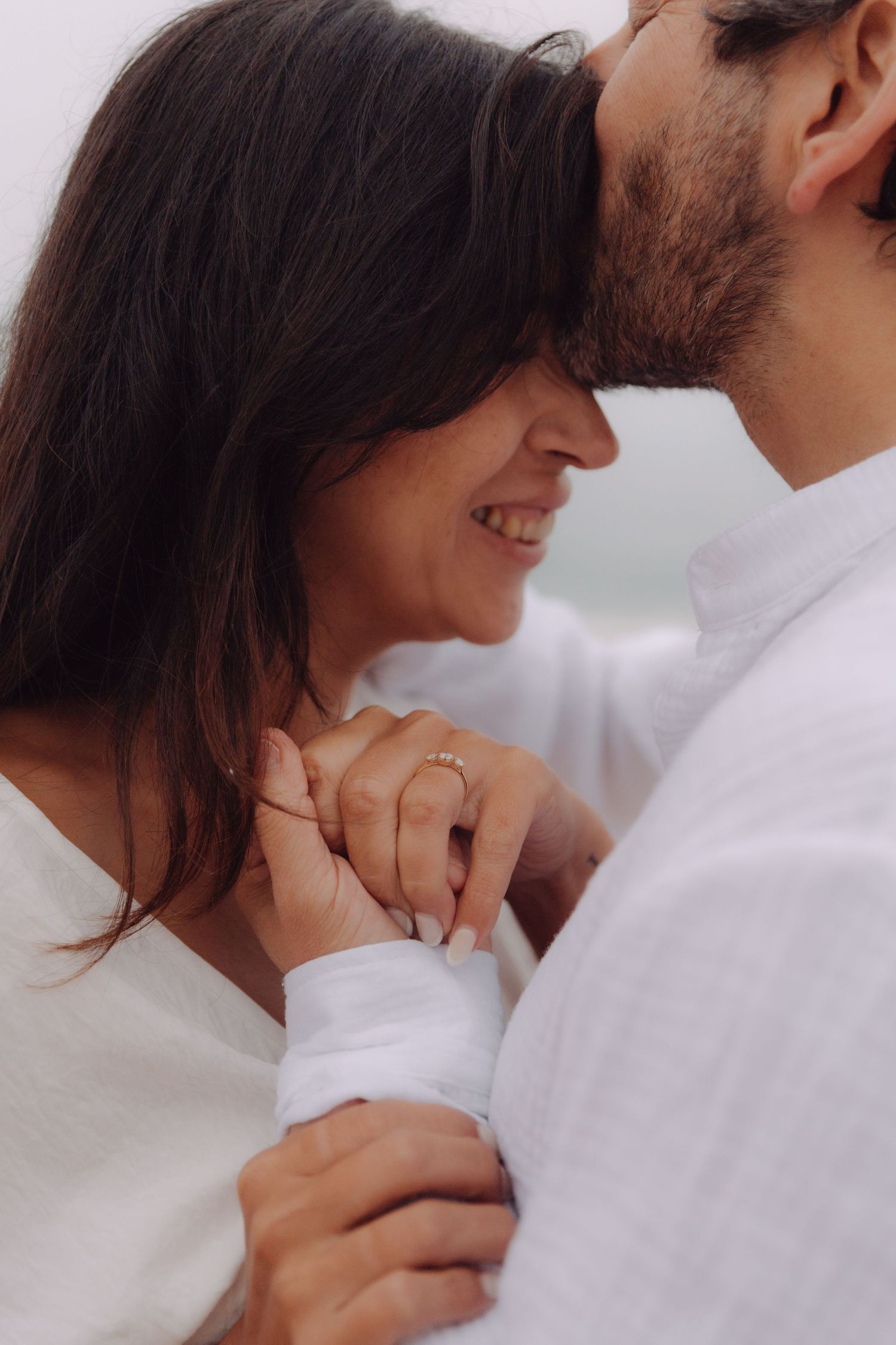 Man kissing smiling woman on forehead while holding hands, engagement ring visible on her finger.