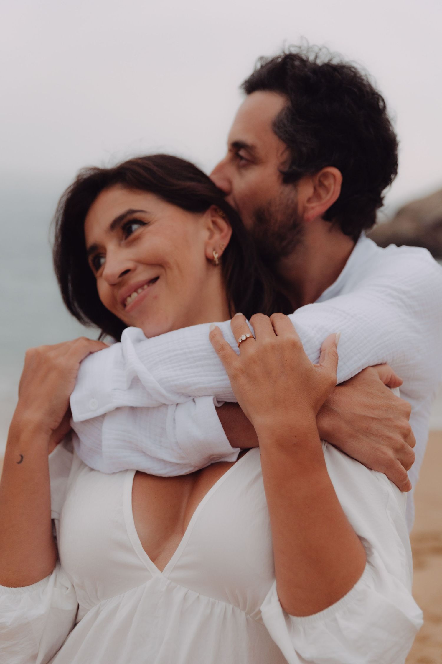 Man embracing woman from behind and kissing her cheek on beach, engagement ring visible on her hand.