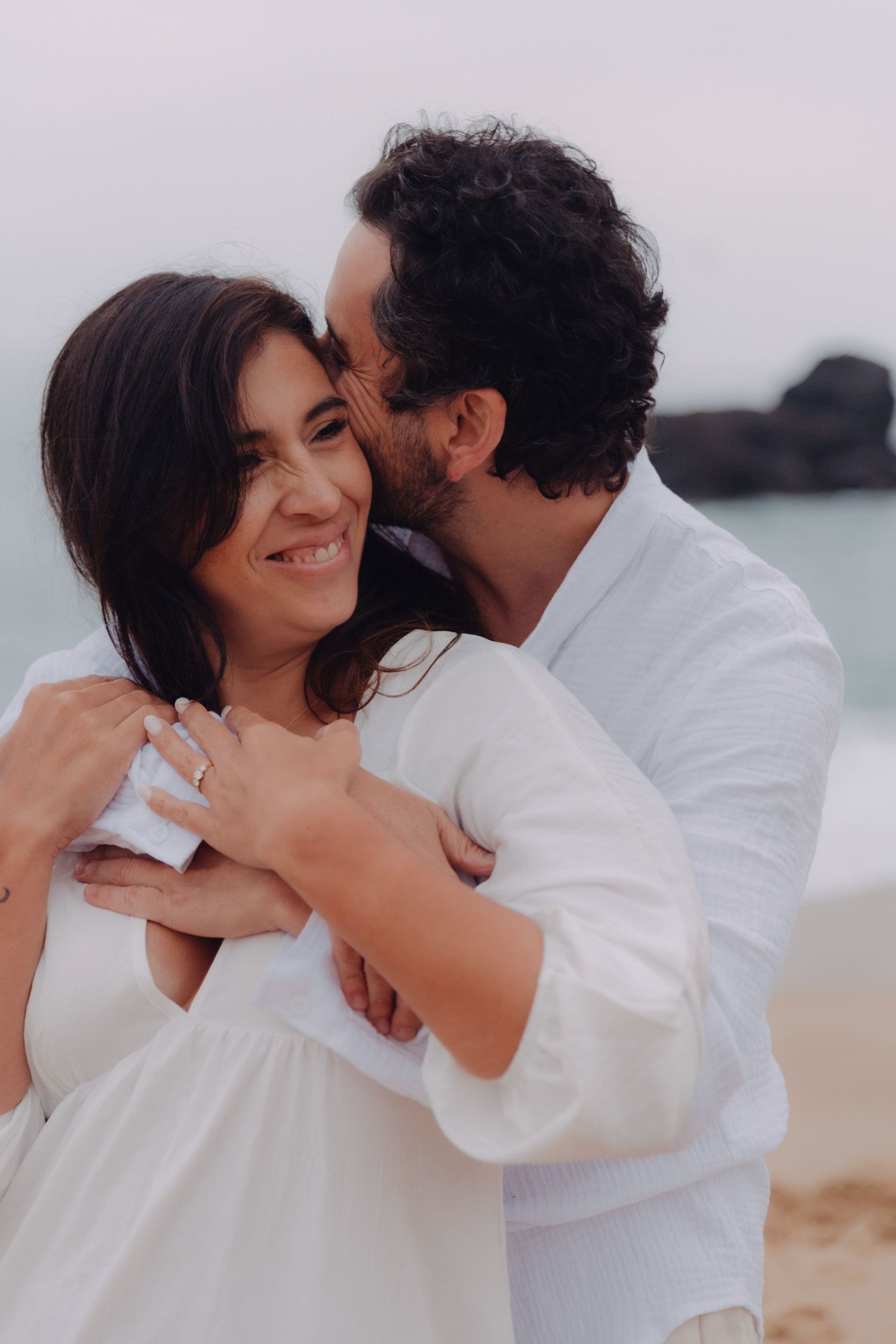 Couple in white on sandy beach, man kissing woman's cheek while she smiles, ring on her hand.