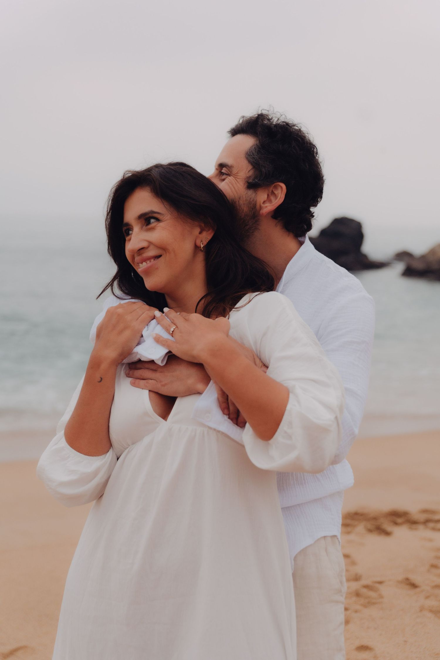 Couple embracing on beach with ocean behind them, woman smiling, man holding her from behind in white.