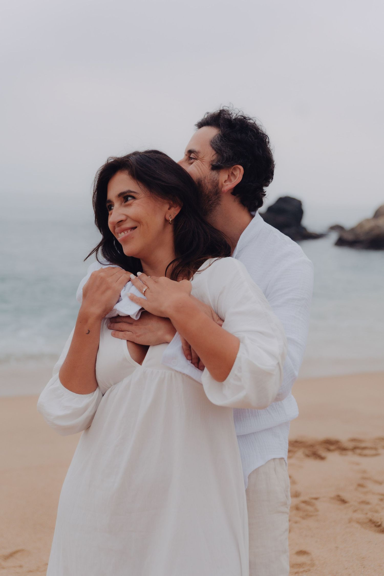 Couple in white linen embracing on sandy beach, woman smiling with ocean and rocks in background.