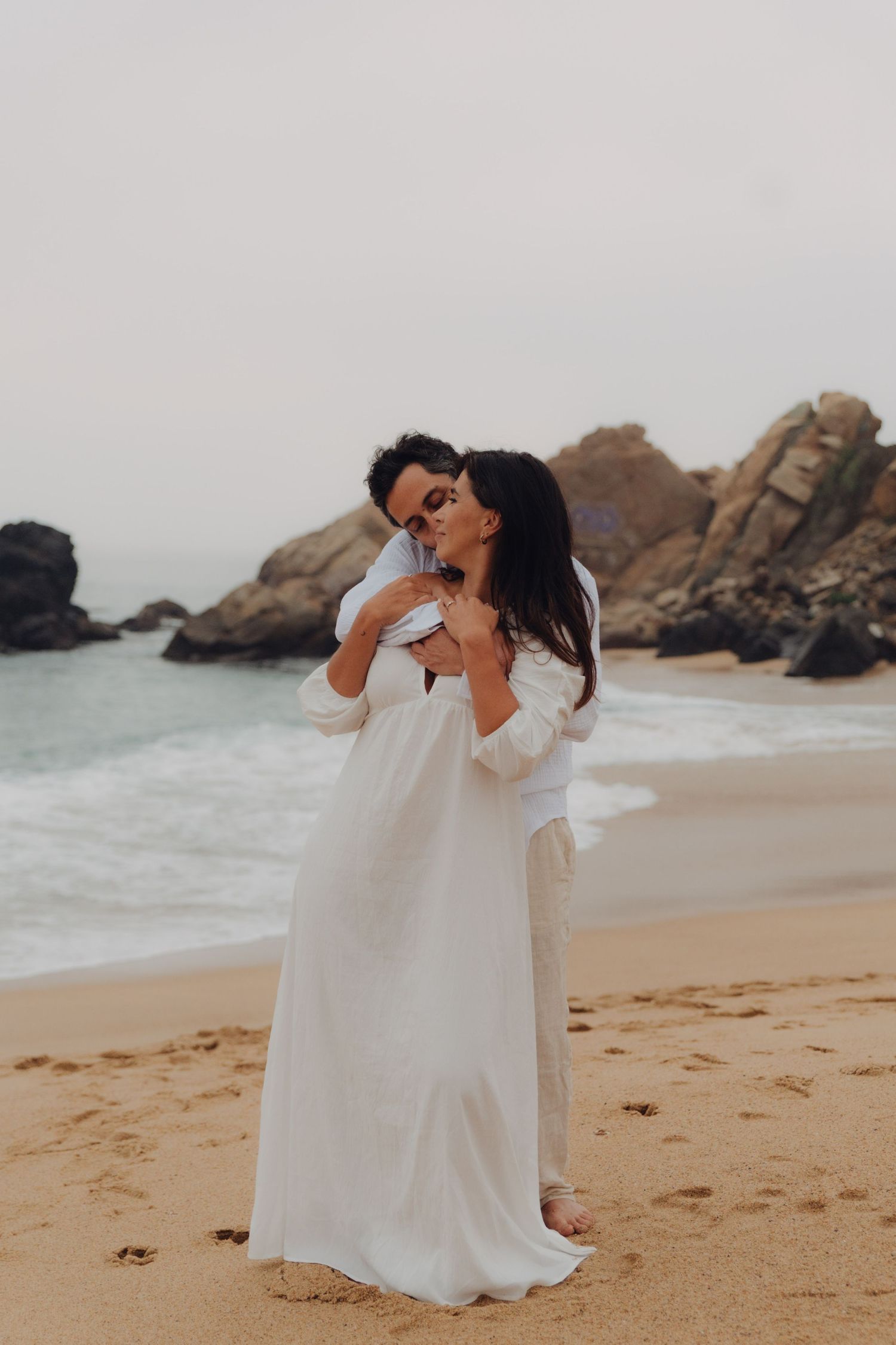 Romantic couple embracing on sandy beach, woman in white gown, man in white shirt, with rocky ocean backdrop.