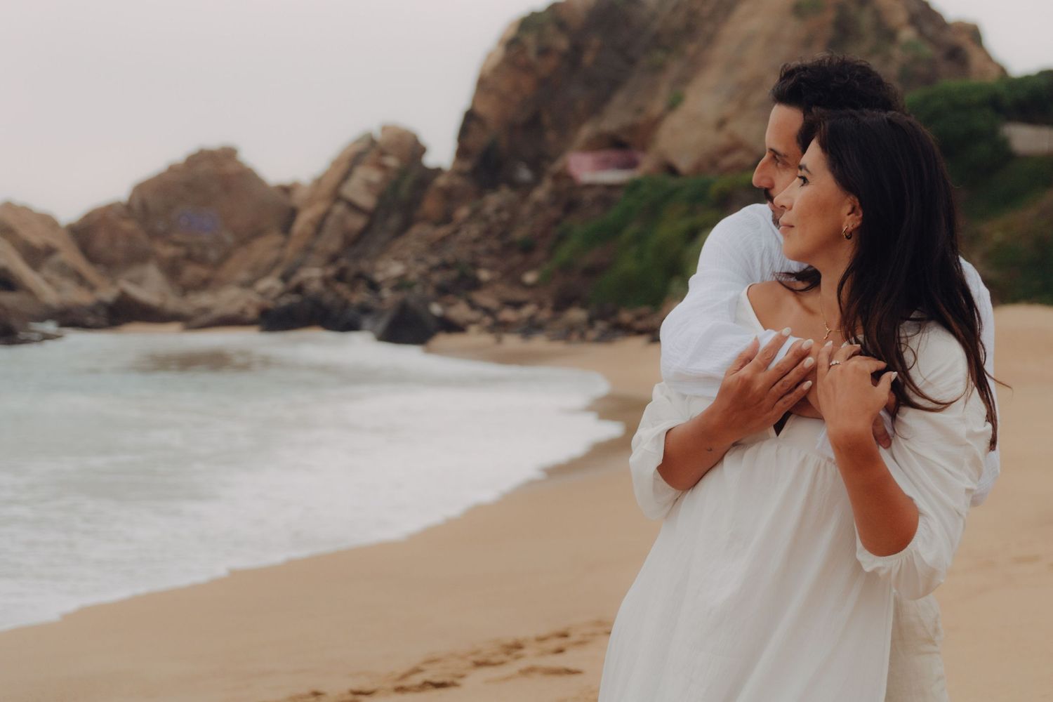 Couple sharing tender moment on beach, man embracing woman in white dress with rocky coastline behind them.