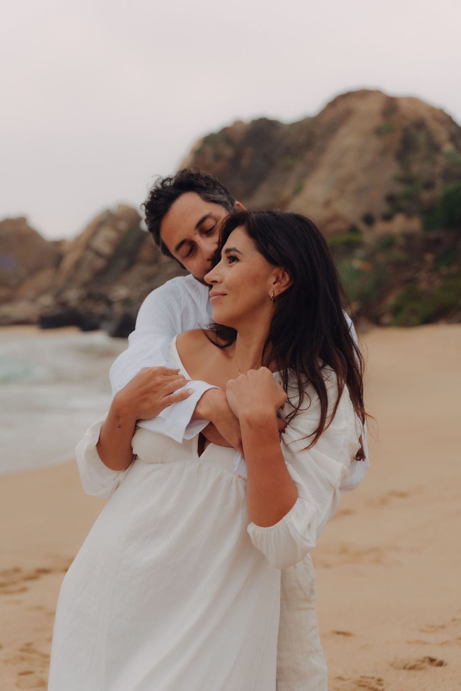 Man tenderly embracing woman in white off-shoulder dress on sandy beach with ocean rocks in background.
