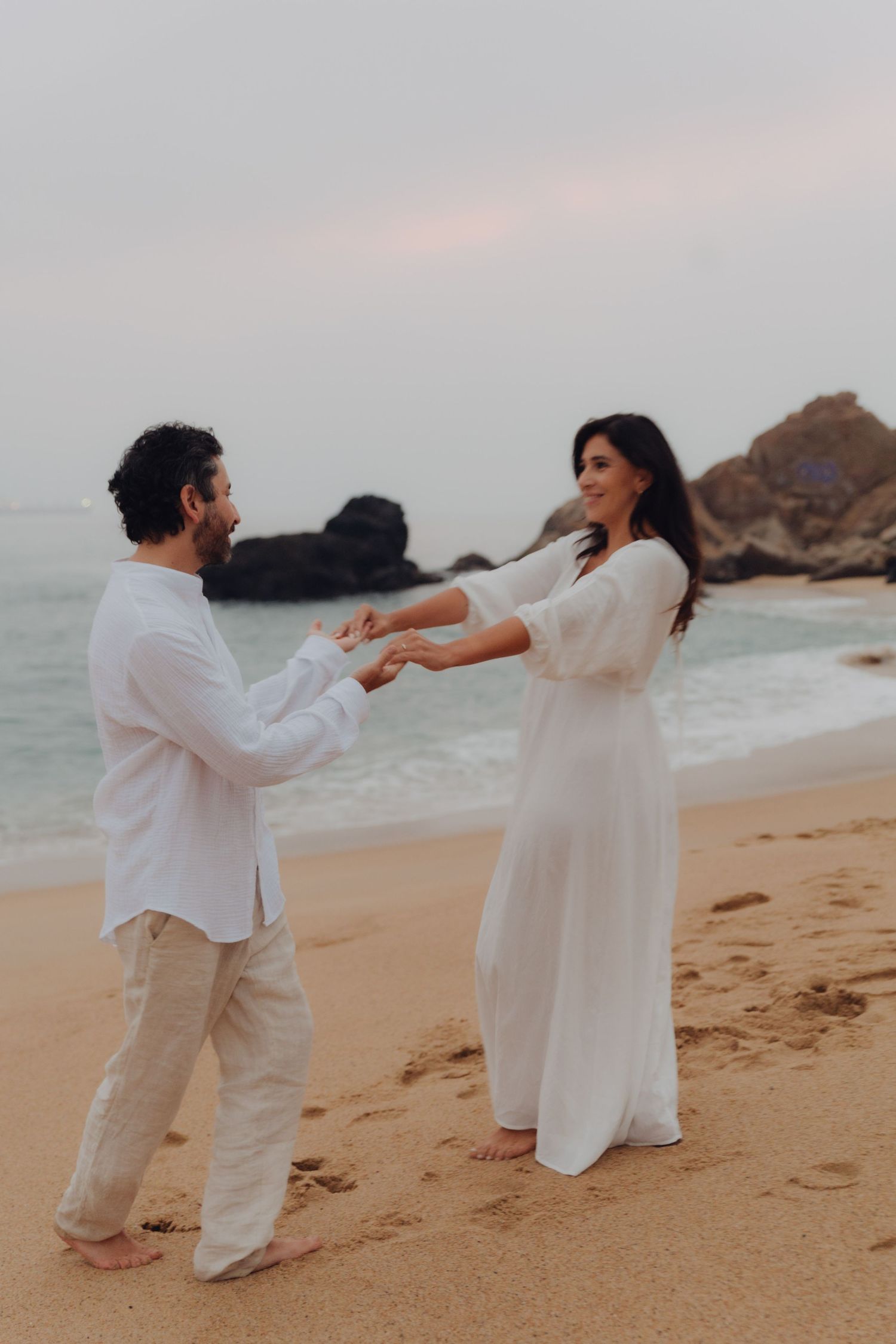 Romantic couple holding hands and dancing on sandy beach at dusk with rocky ocean shore behind them.