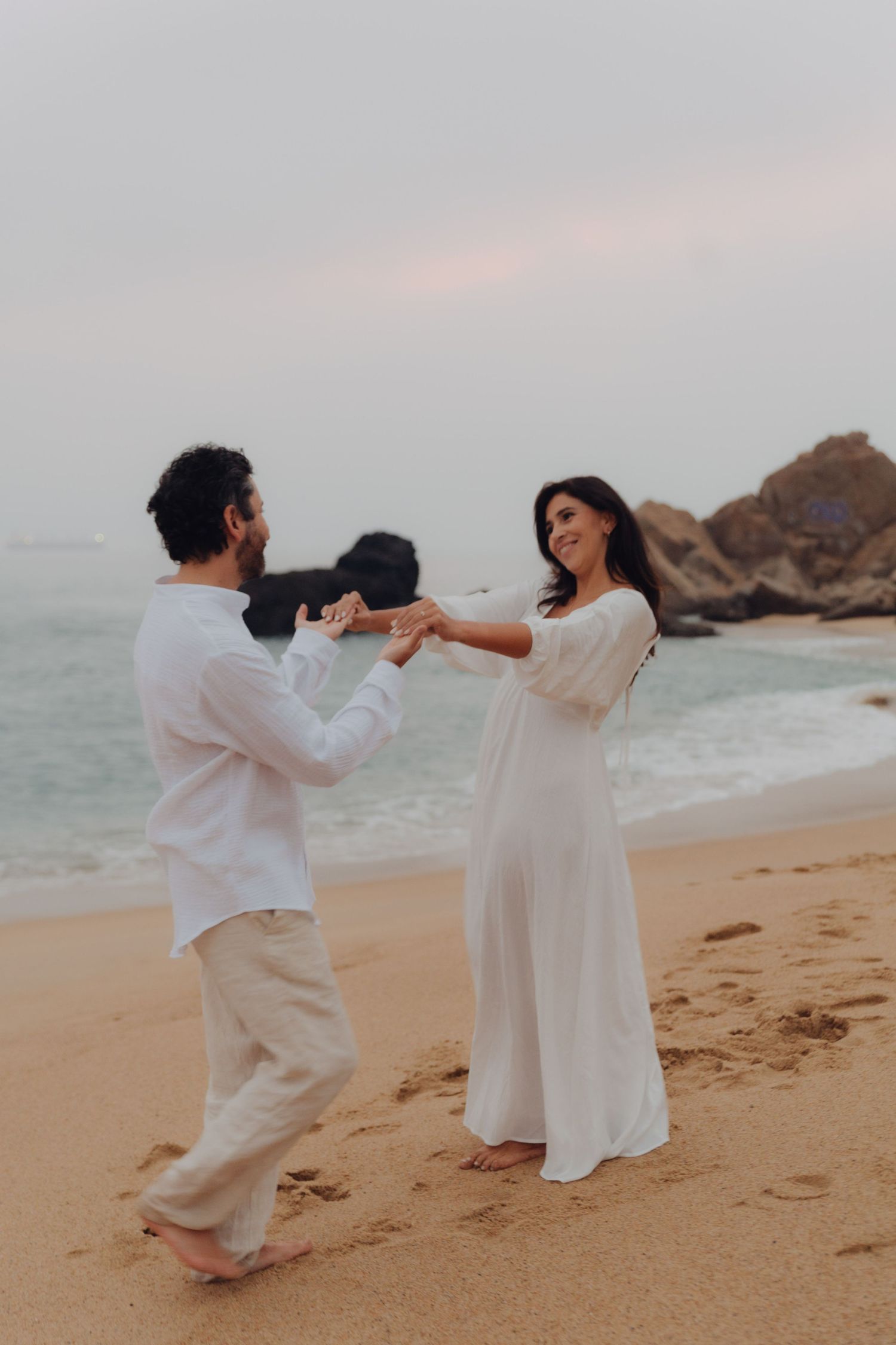 Smiling couple in white outfits holding hands playfully on sandy beach with ocean waves and rocks behind.
