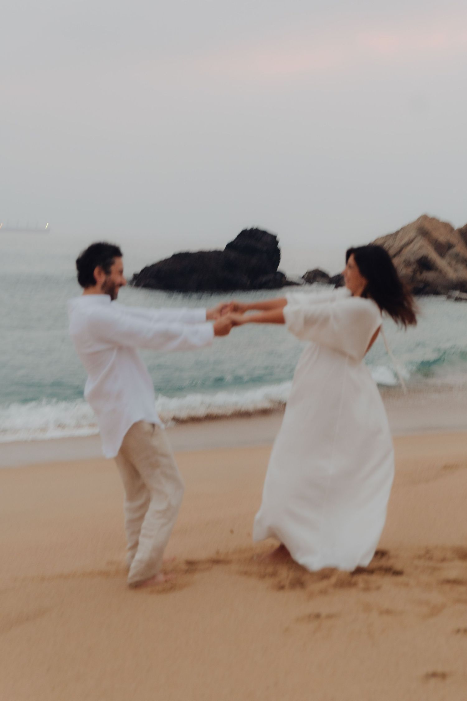 Couple spinning and dancing together on sandy beach at dusk with dramatic ocean rocks in background.