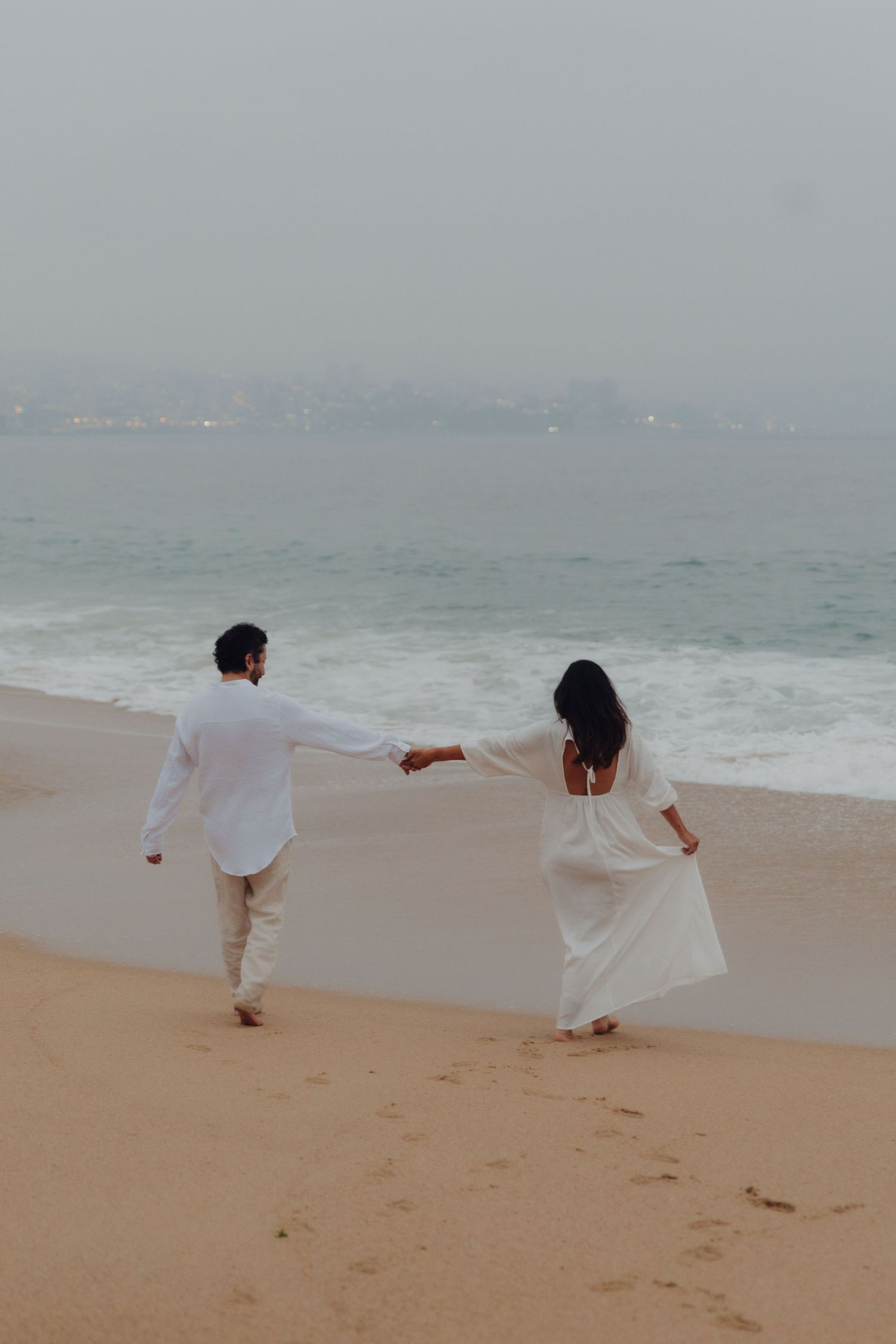 Couple in white outfits walking hand in hand along misty sandy beach toward the ocean horizon.