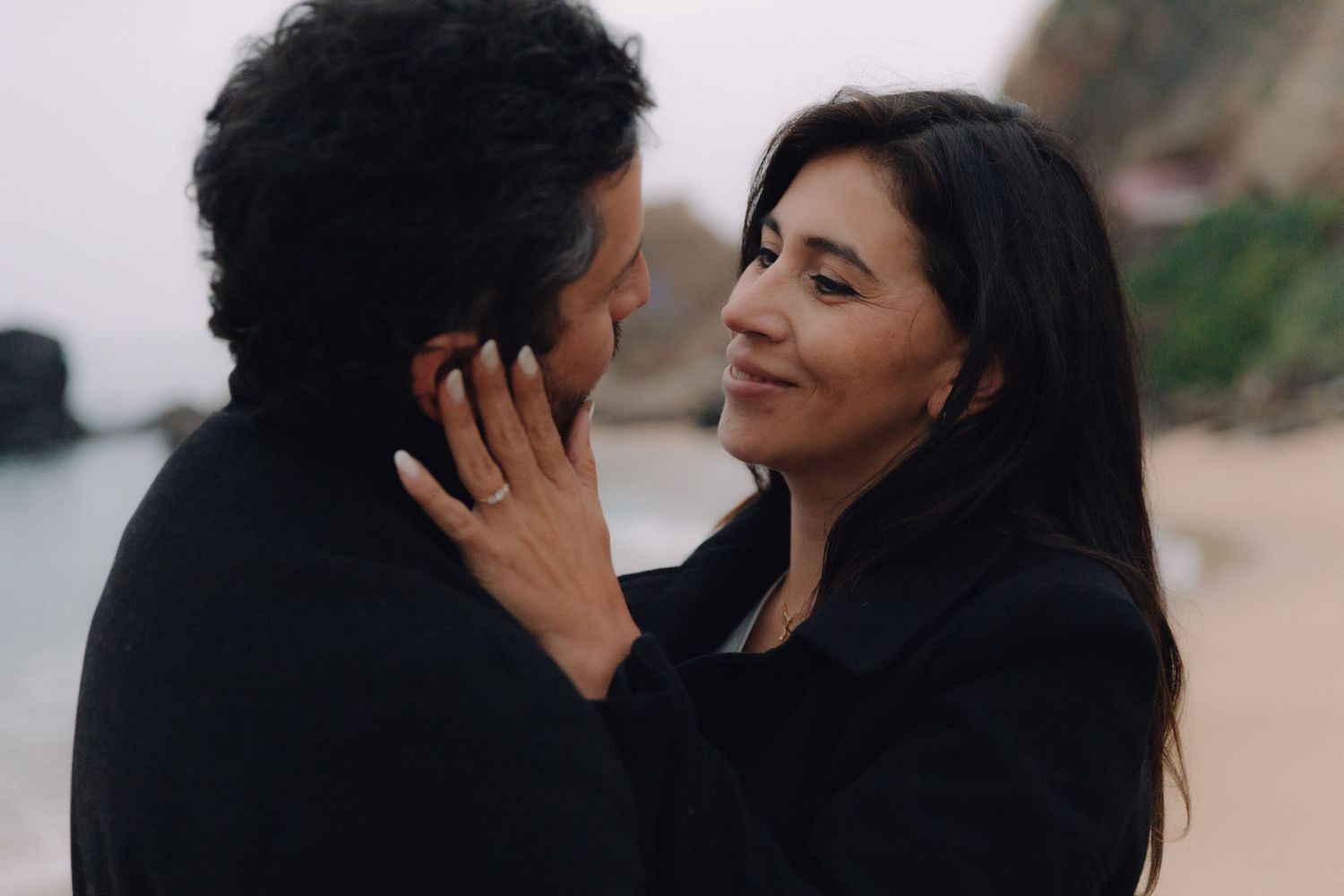 Couple in black coats sharing intimate moment on beach, woman smiling tenderly at man with ocean behind.