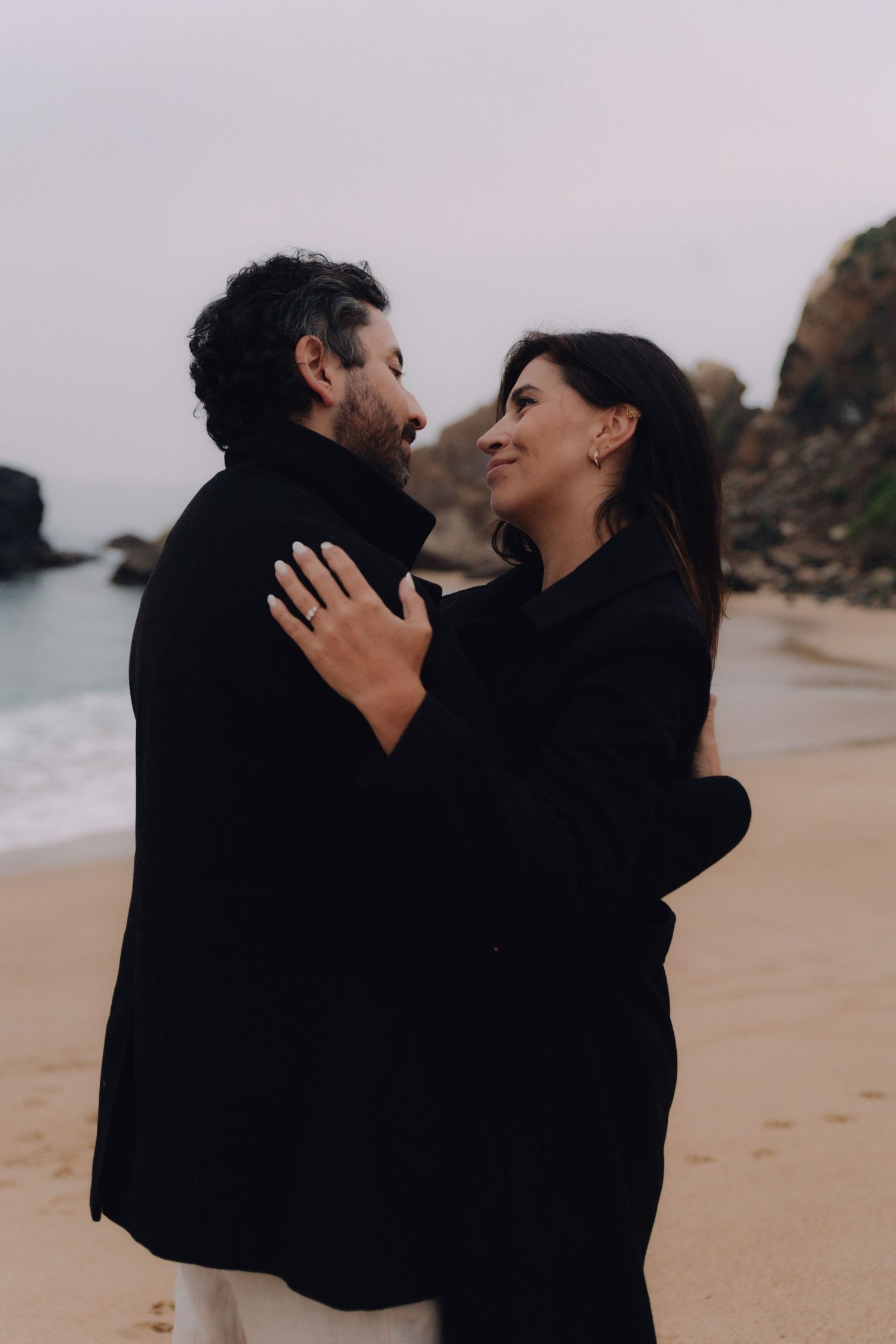 Romantic couple in matching black coats embracing on sandy beach with rocky coastline in the background.