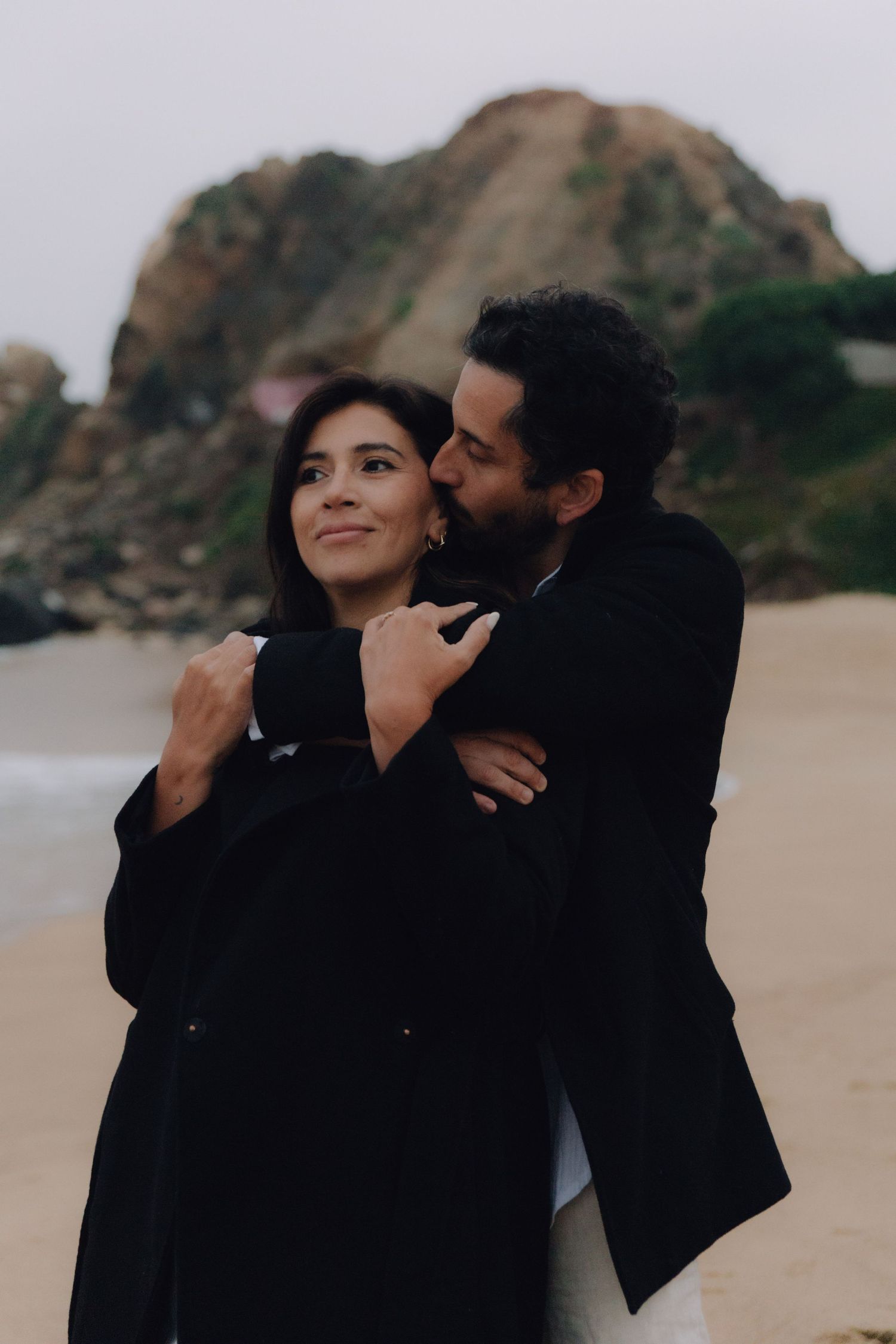 Man in black coat kissing woman on cheek while embracing her on sandy beach with rocky cliffs behind.