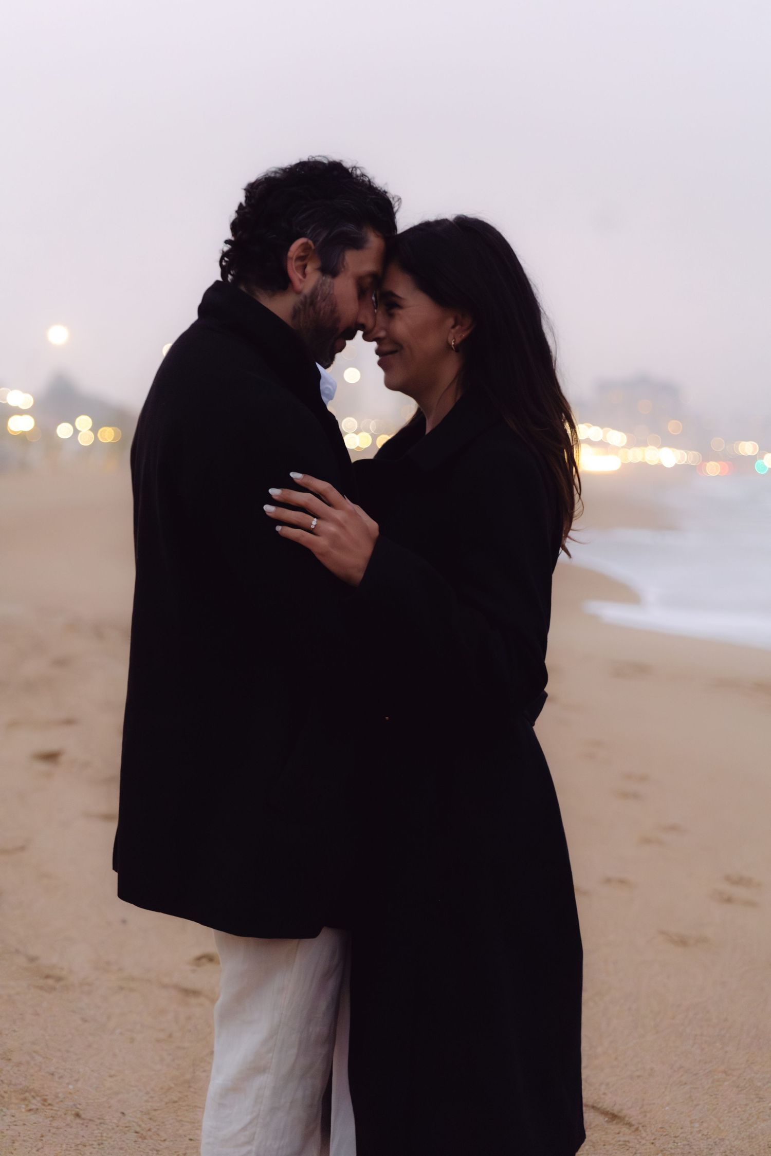 Romantic couple embracing on a beach at dusk, wearing black coats with bokeh city lights in the background.