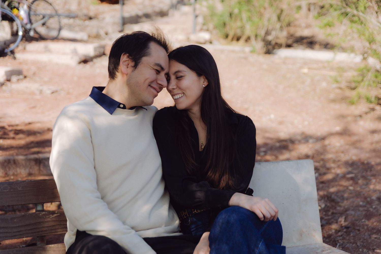 Affectionate couple leaning foreheads together and smiling while seated on an outdoor bench.
