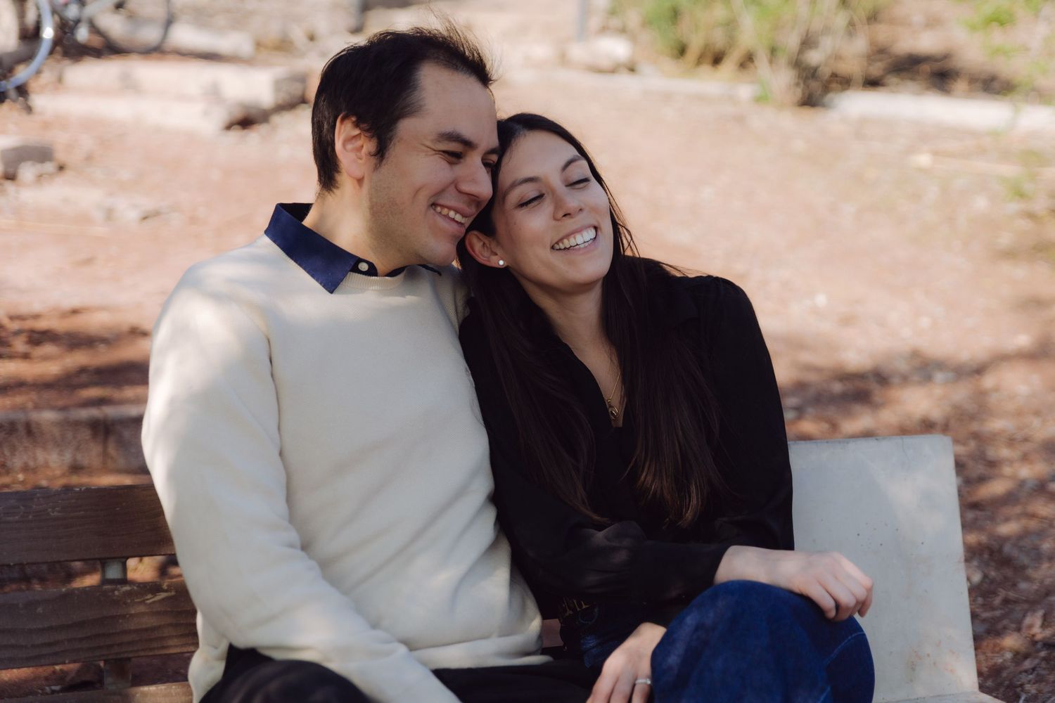 Happy couple laughing together while sitting on a wooden bench in an outdoor park setting.