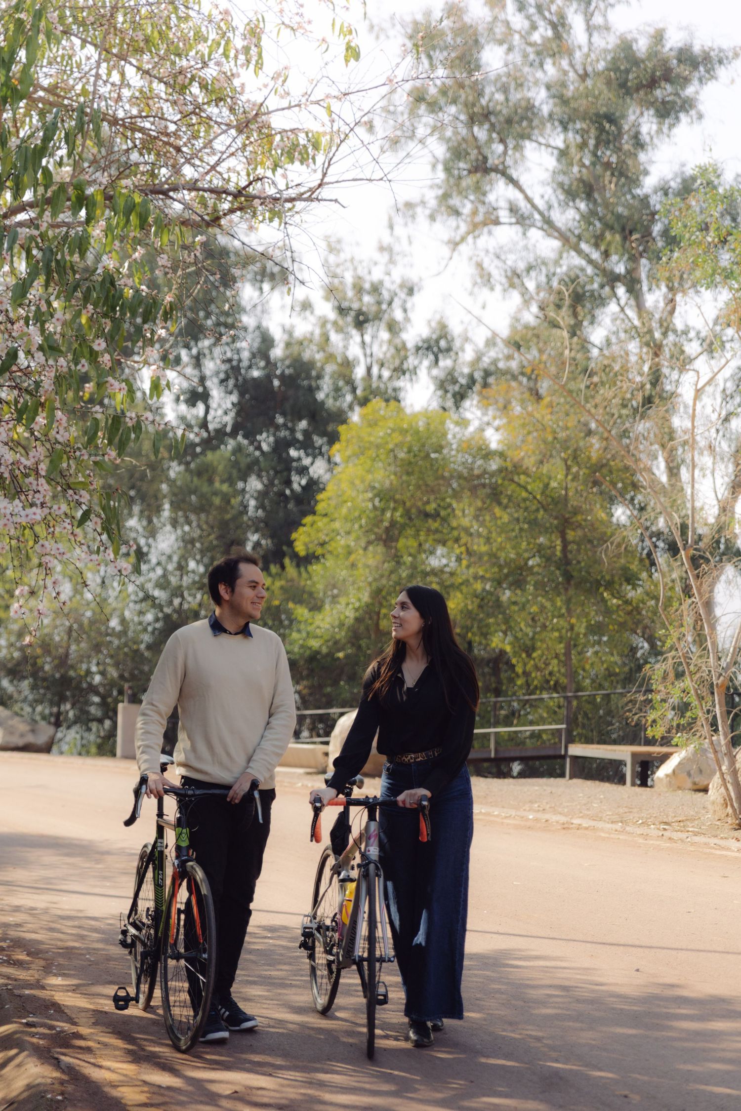 Smiling couple strolling with bicycles on a sunlit path in a green park.