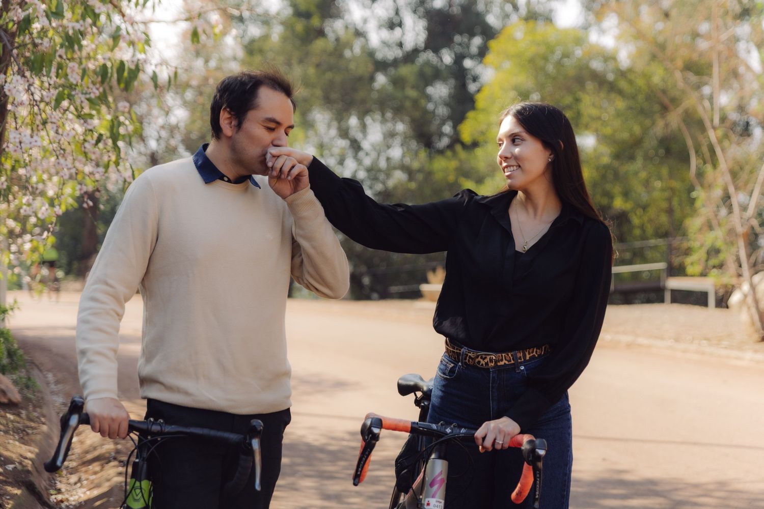 Man kissing woman's hand while holding bikes on a tree-lined park path.